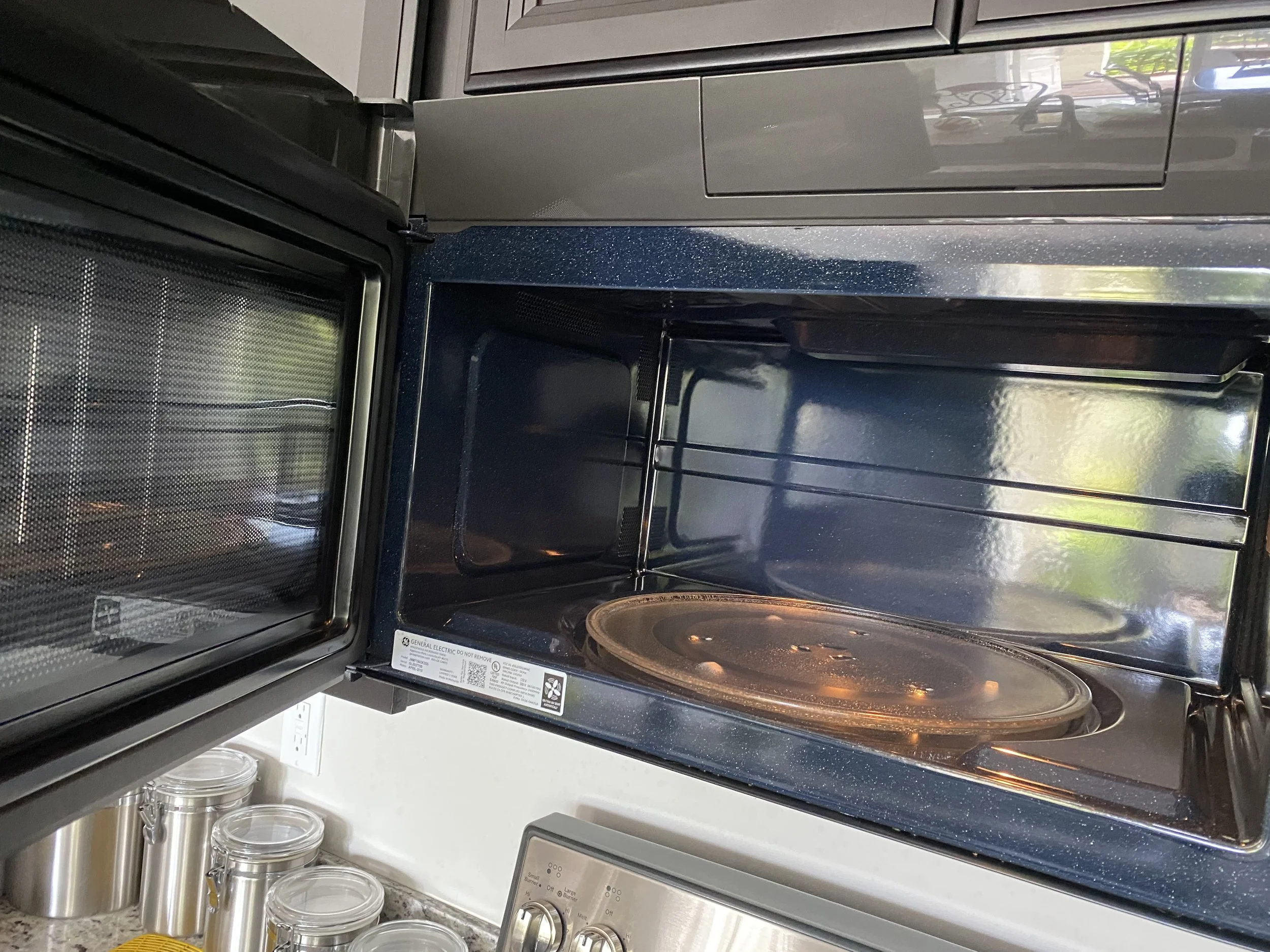 An empty stainless steel microwave oven with its door open, situated on a kitchen countertop next to glass jars and containers.