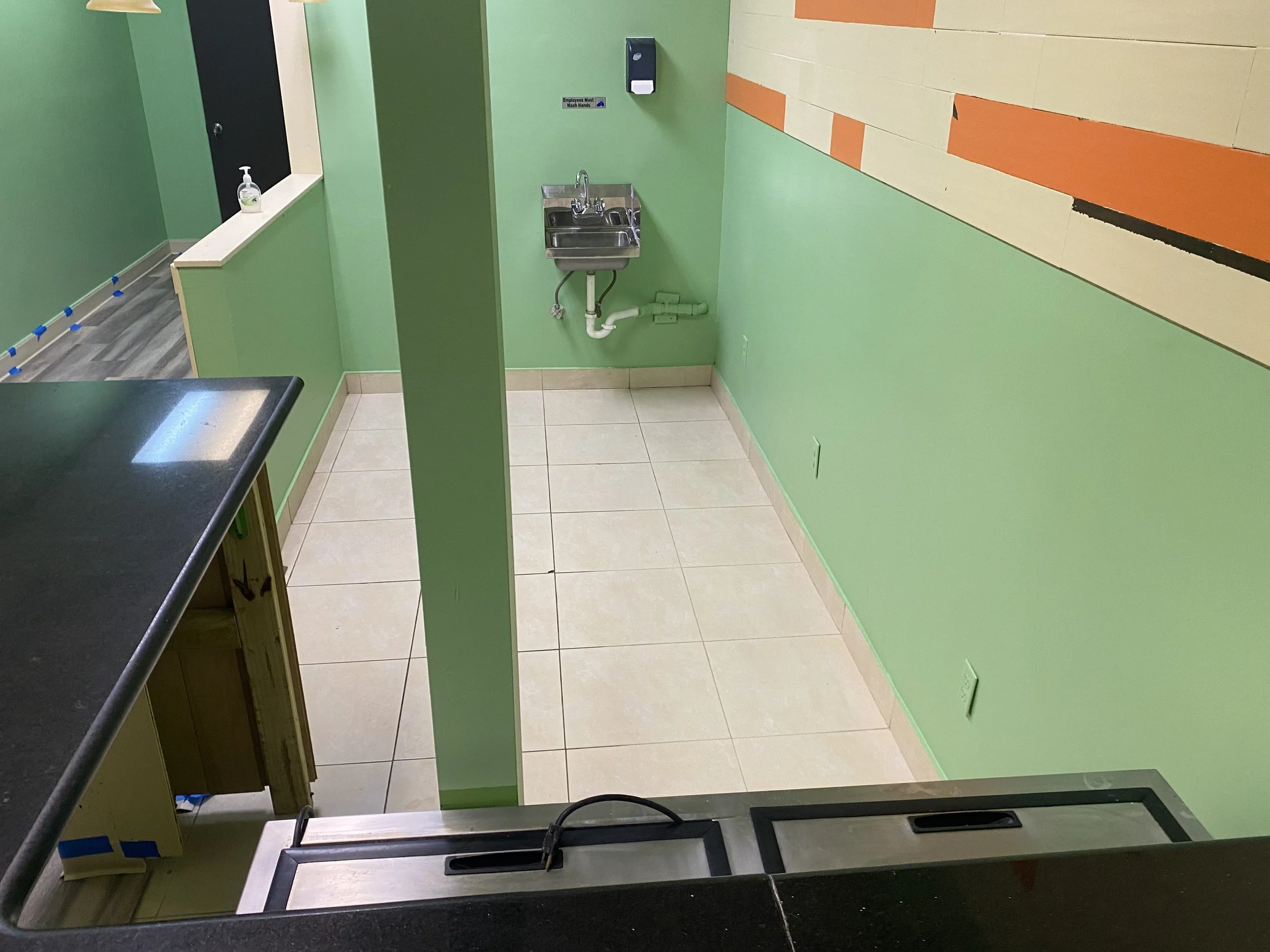 Restaurant Cleaning, A small stainless steel sink with a faucet mounted on a green wall, with a soap dispenser above it and a sign indicating hand washing requirements nearby. Part of countertop and a black object are visible in the foreground.