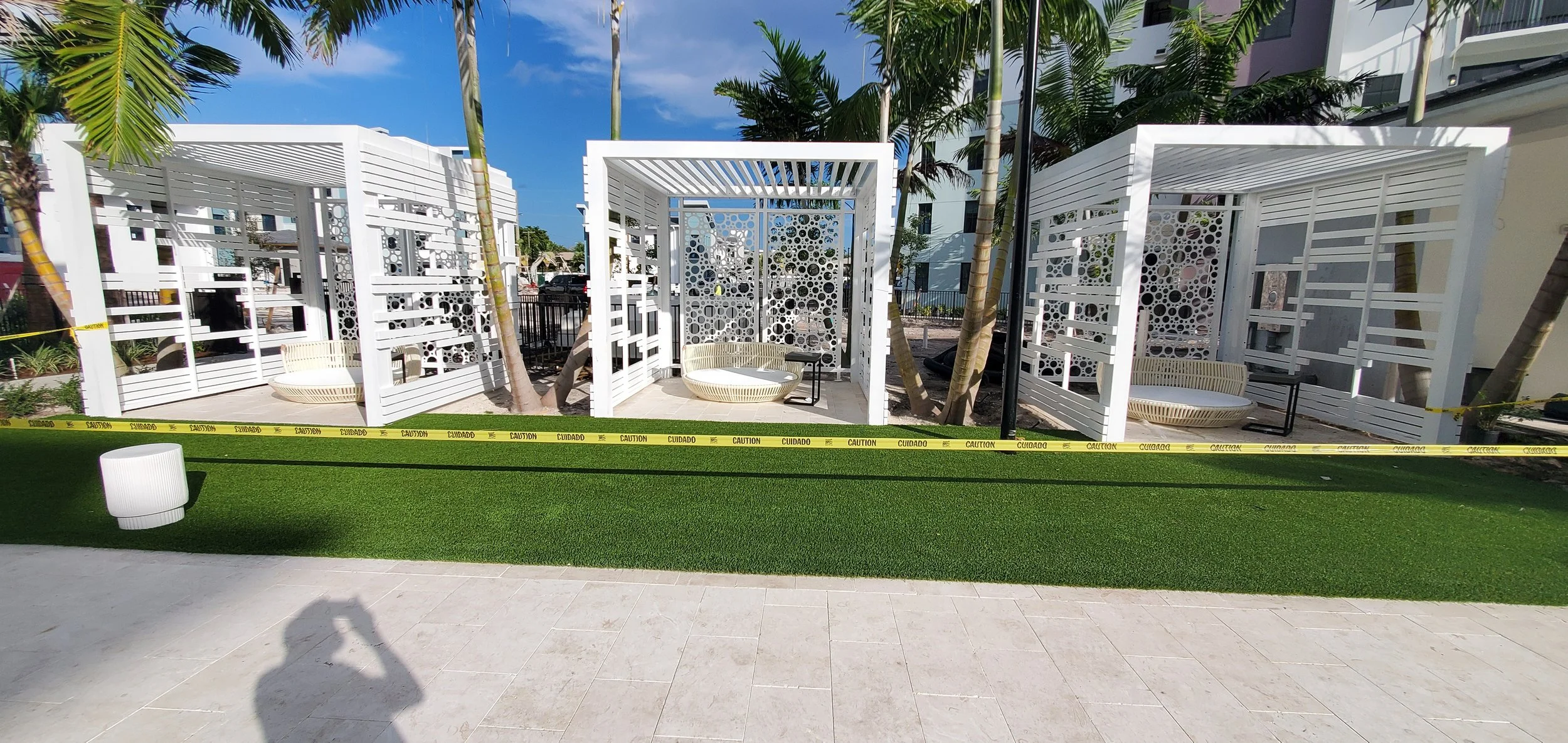White modern outdoor cabanas with circular patterns, surrounded by palm trees, on a sunny day with a clear blue sky, with caution tape in the foreground.