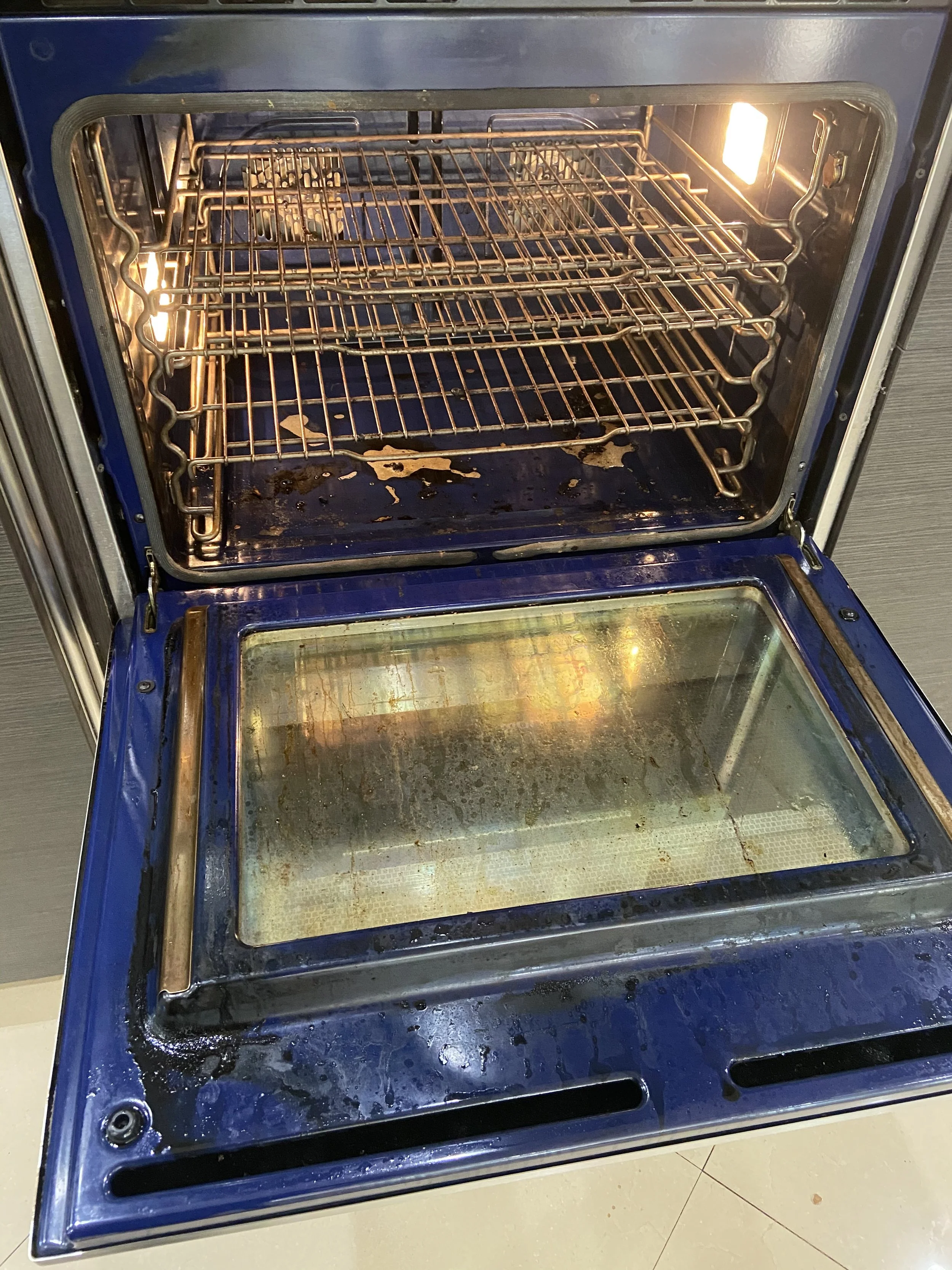 An electric oven with an open door showing a clean interior and empty racks, with some grease and stains at the bottom, and a warm light inside.