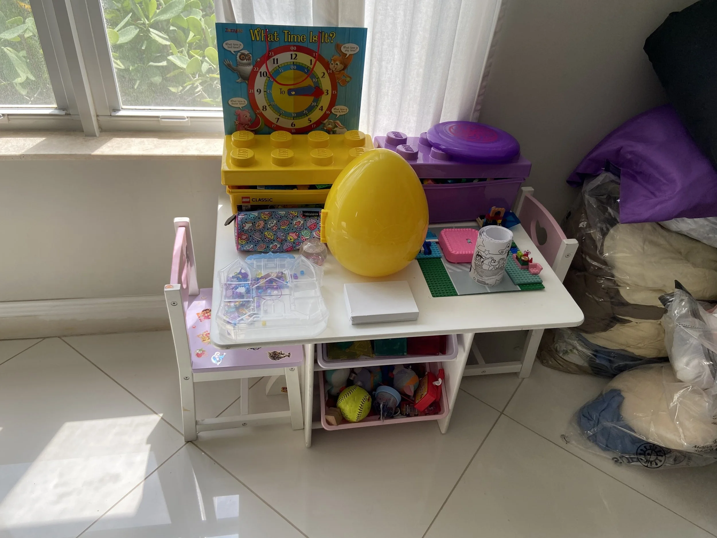 A small white shelf with toys and items including a yellow balloon, a purple plastic container, building blocks, a plastic cup with coloring, a yellow ball, and a Lego display, positioned near a window with curtains. A colorful educational clock and 