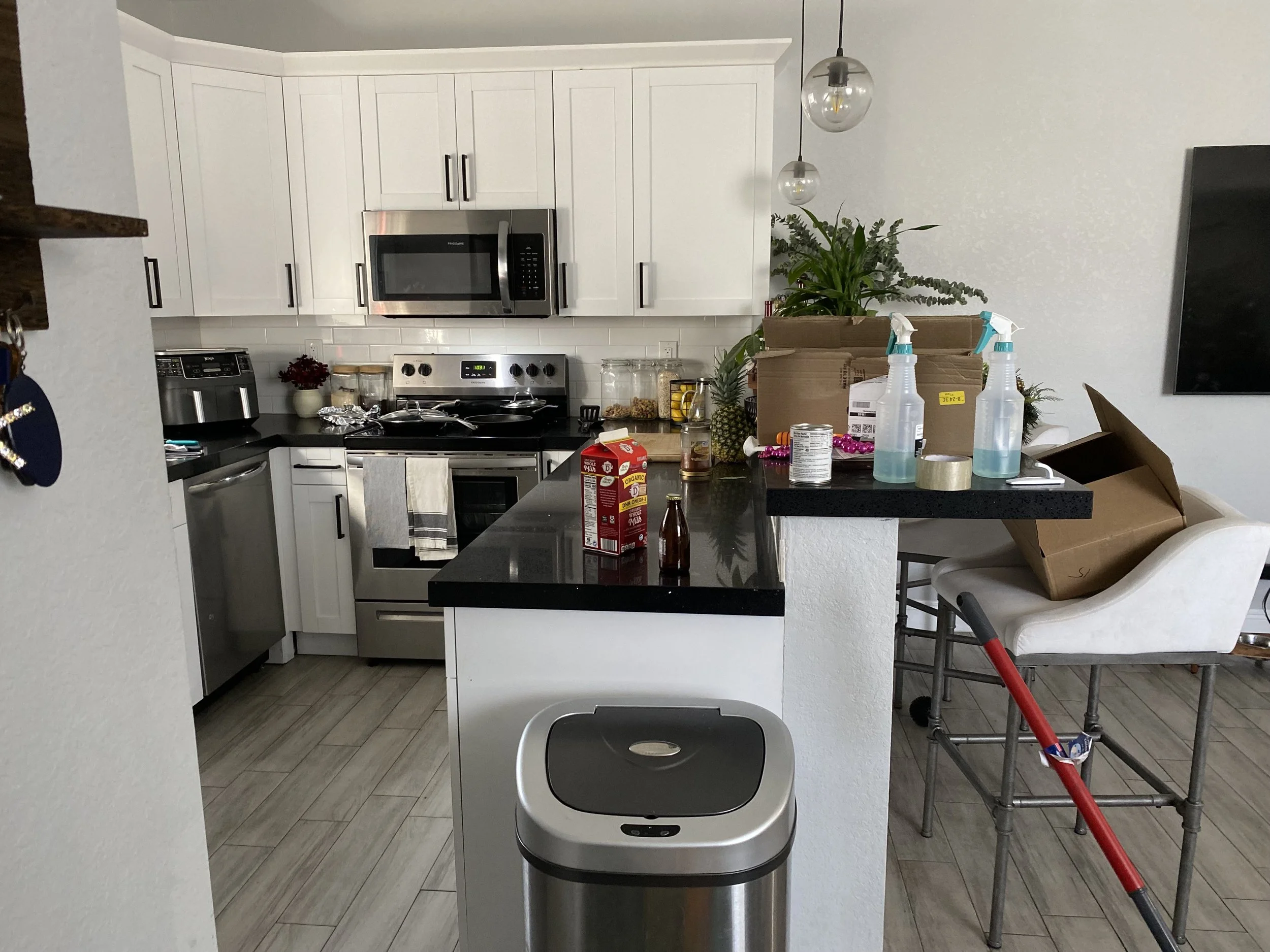 A modern kitchen with white cabinetry, a black countertop, a stainless steel microwave, stove, and dishwasher. The countertop has various items including a pineapple, jars, and I also see three cleaning spray bottles, a cardboard box, and some snacks