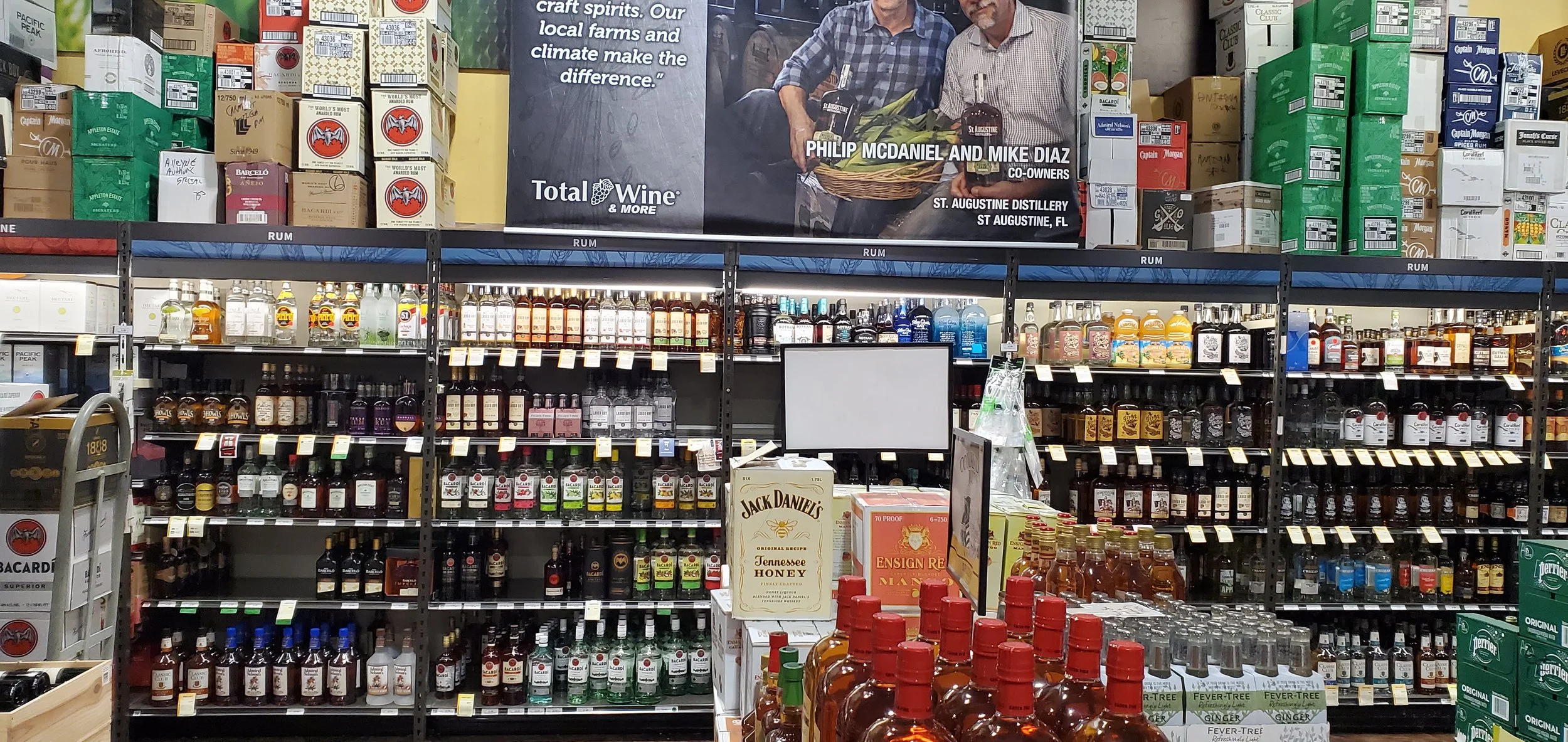 A retail store shelf stocked with various bottles of alcohol, including rum, whiskey, and bourbon, with a large promotional banner above advertising local farms and distilleries, featuring two men holding a basket of produce.