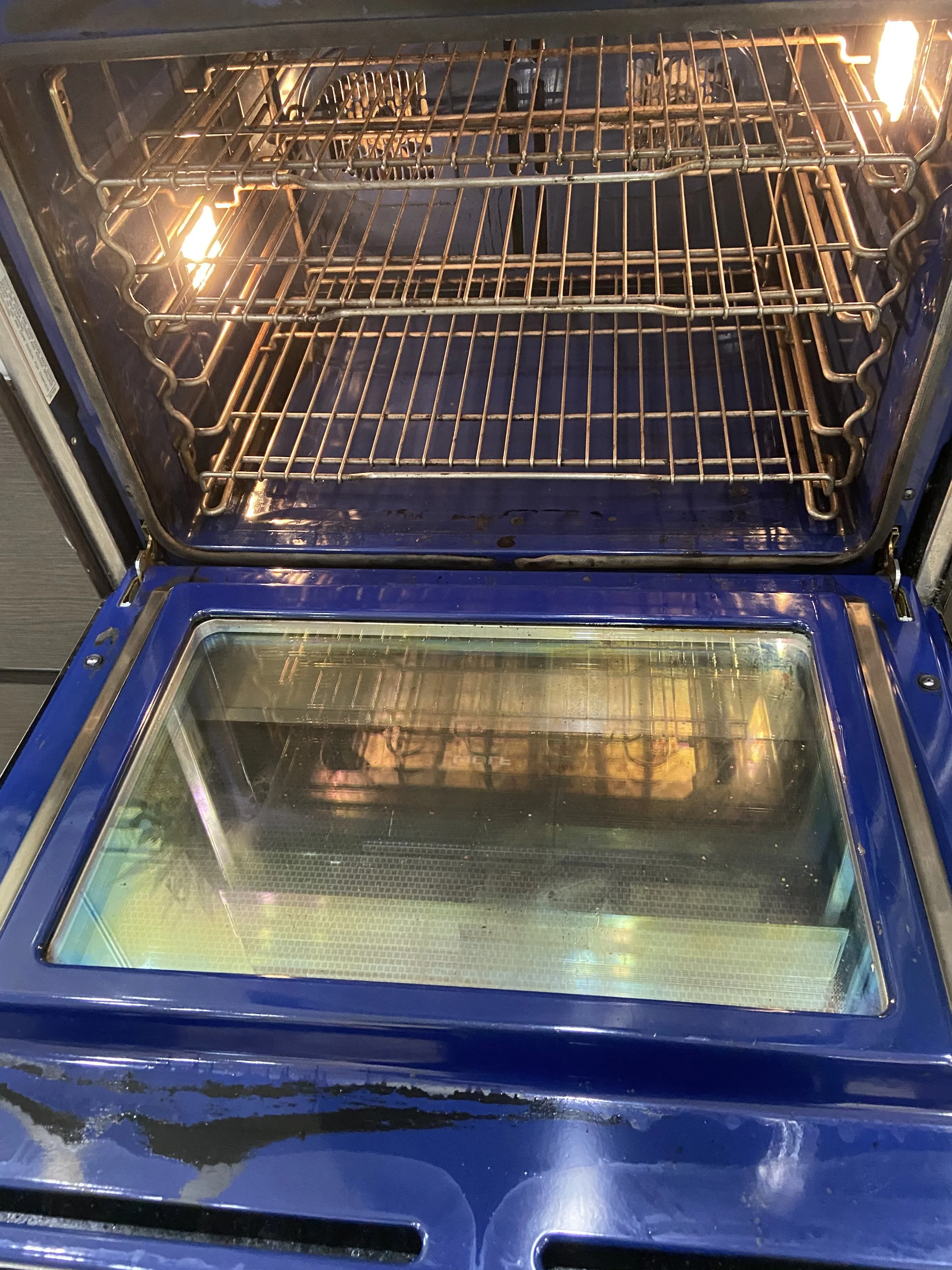 An open oven with two racks inside, a blue exterior, and a glass door. The oven appears empty and turned on, with a light illuminating the interior.