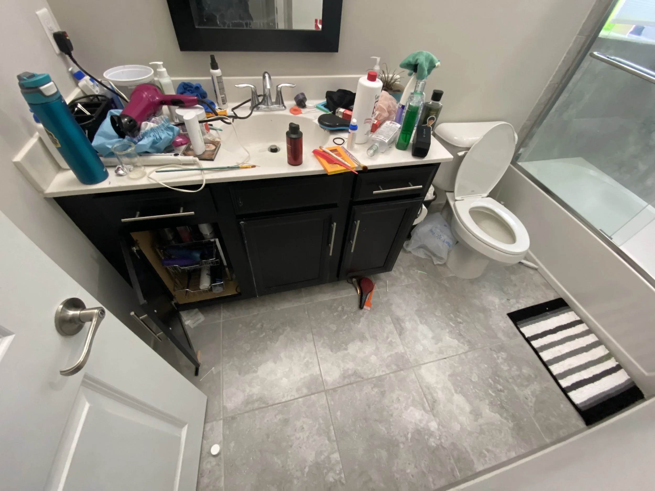 Bathroom sink and countertop cluttered with various personal care items, a toilet with the lid up, and a shower door with a striped rug on the floor.