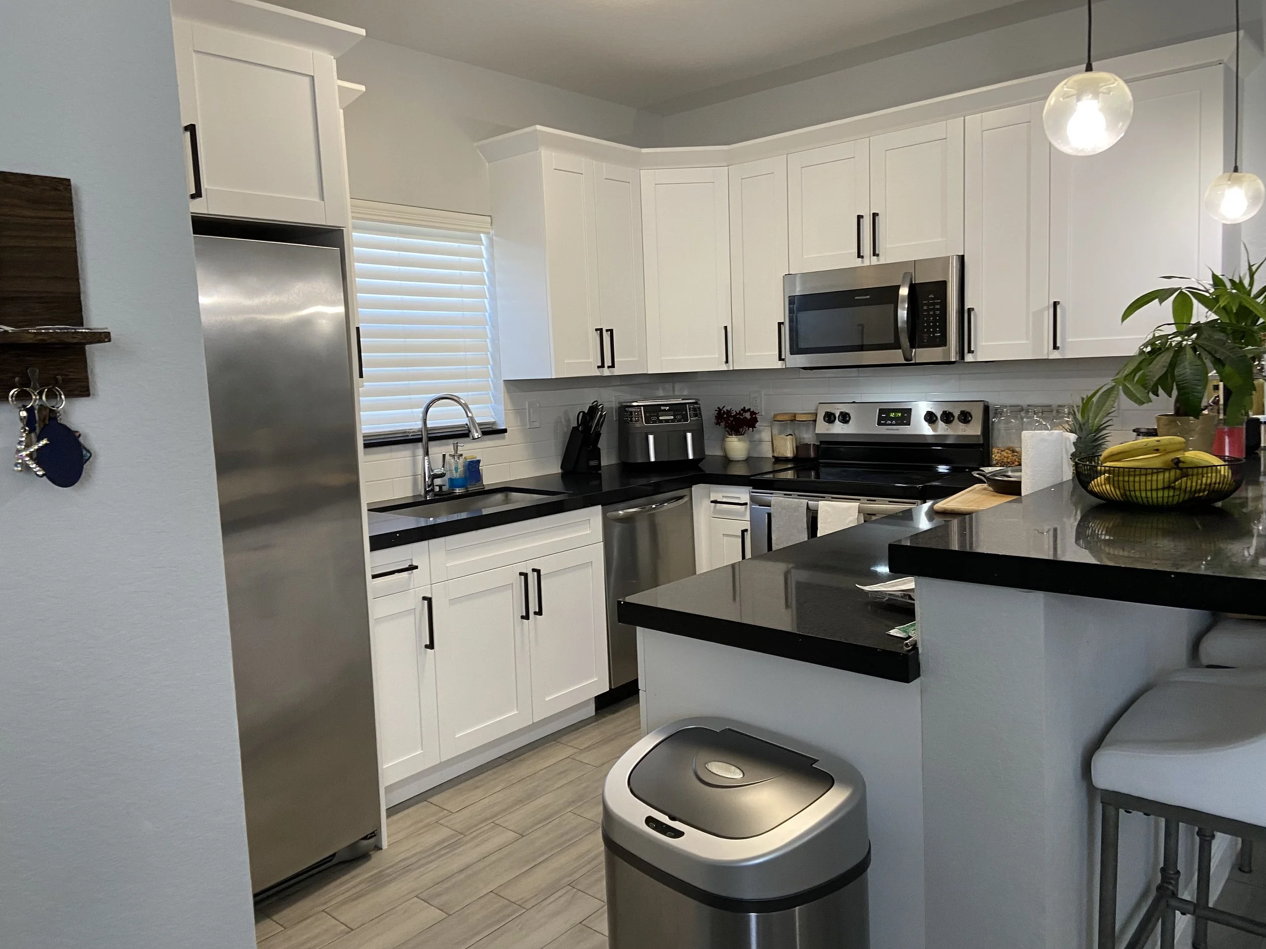 A modern kitchen with white cabinets, stainless steel appliances, a black countertop, and a gray trash bin. There is a window with blinds, a potted plant, and hanging lights.