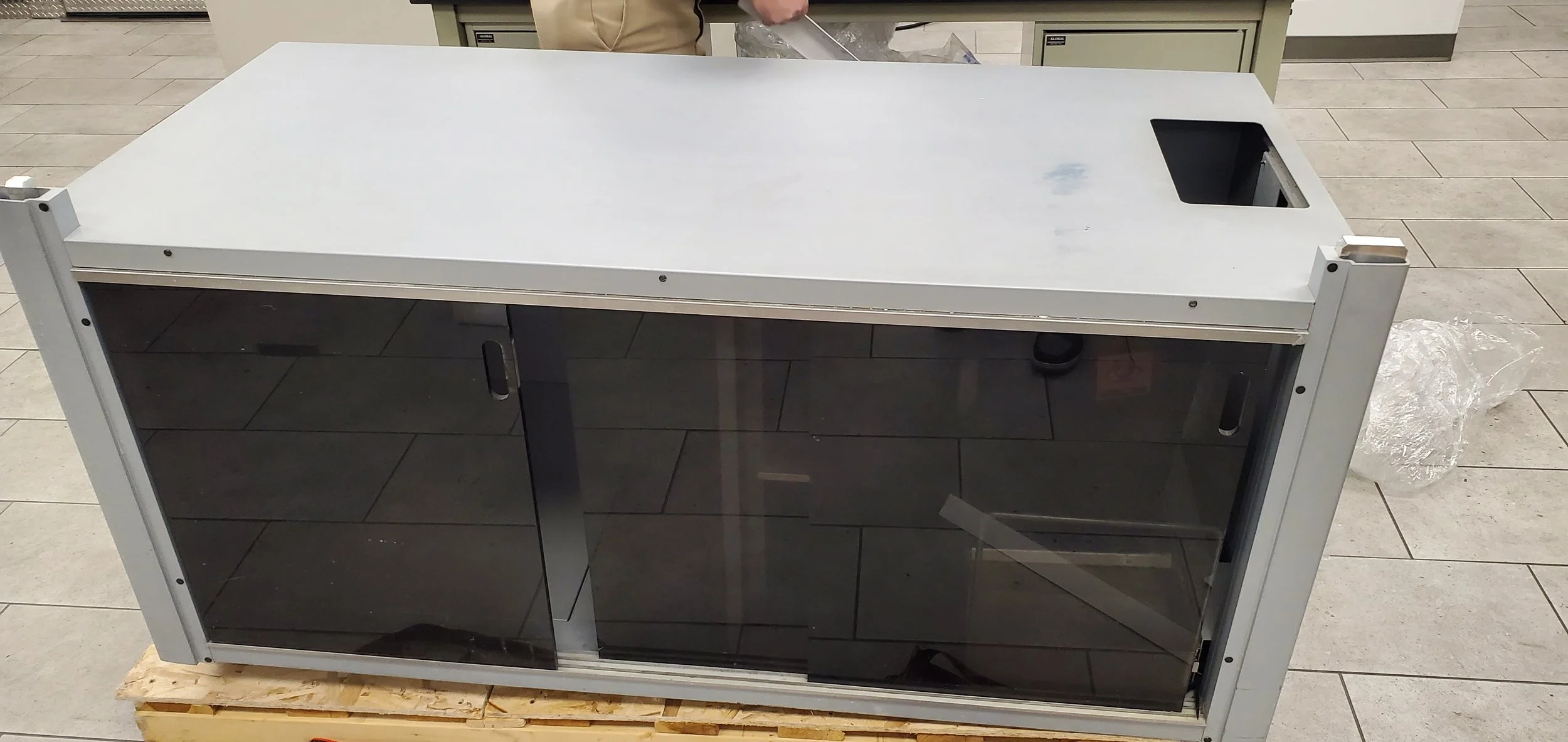 Installation Hollywood, Pembroke Pines, Coral Springs, Miramar Lauderhill, A metallic kitchen or laboratory appliance with a built-in sink on the right, glass doors, and sliding handles, placed on a wooden pallet in a retail or storage setting.