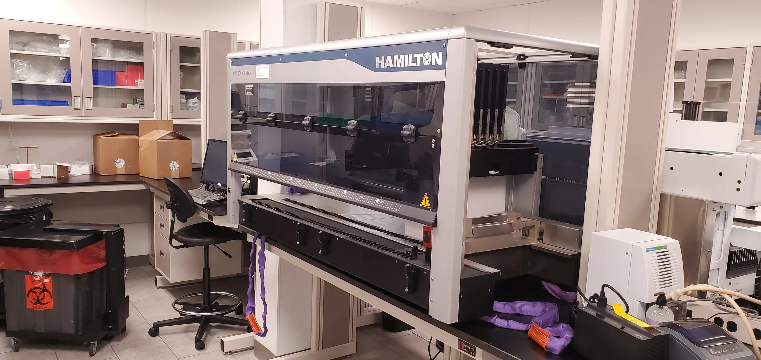 Laboratory room with scientific equipment, including a Hamilton robotic system, cabinets with supplies, a computer, boxes, and a trash bin.