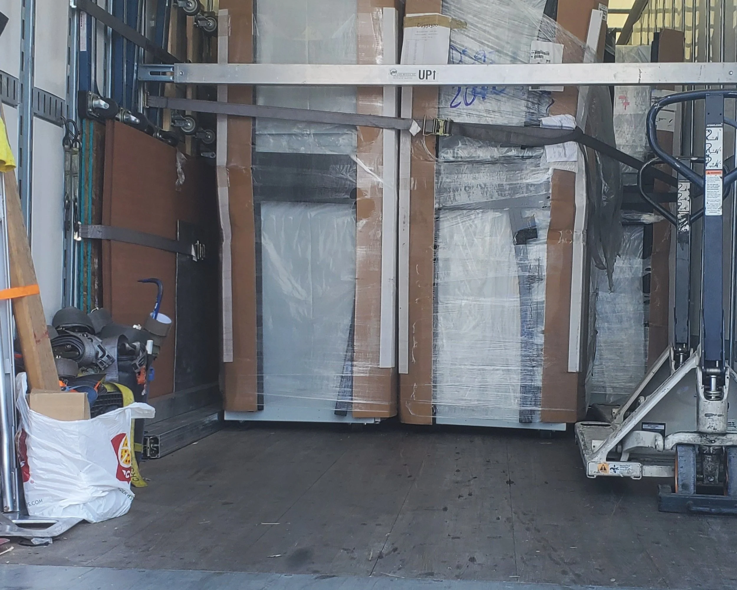 Professional pickup and delivery services, A warehouse or storage room with a pallet jack, cardboard boxes, plastic-wrapped pallets, and various tools and equipment stored on shelves.