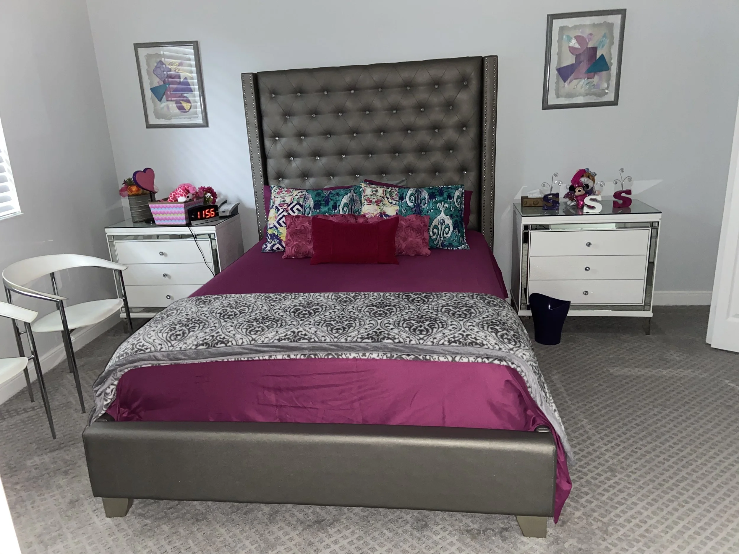 A bedroom with a large bed and a tall, tufted headboard, decorated with colorful pillows and a maroon bedspread. There are two white nightstands on each side, with decorative items and a digital clock. Two framed abstract art pieces hang on the wall 