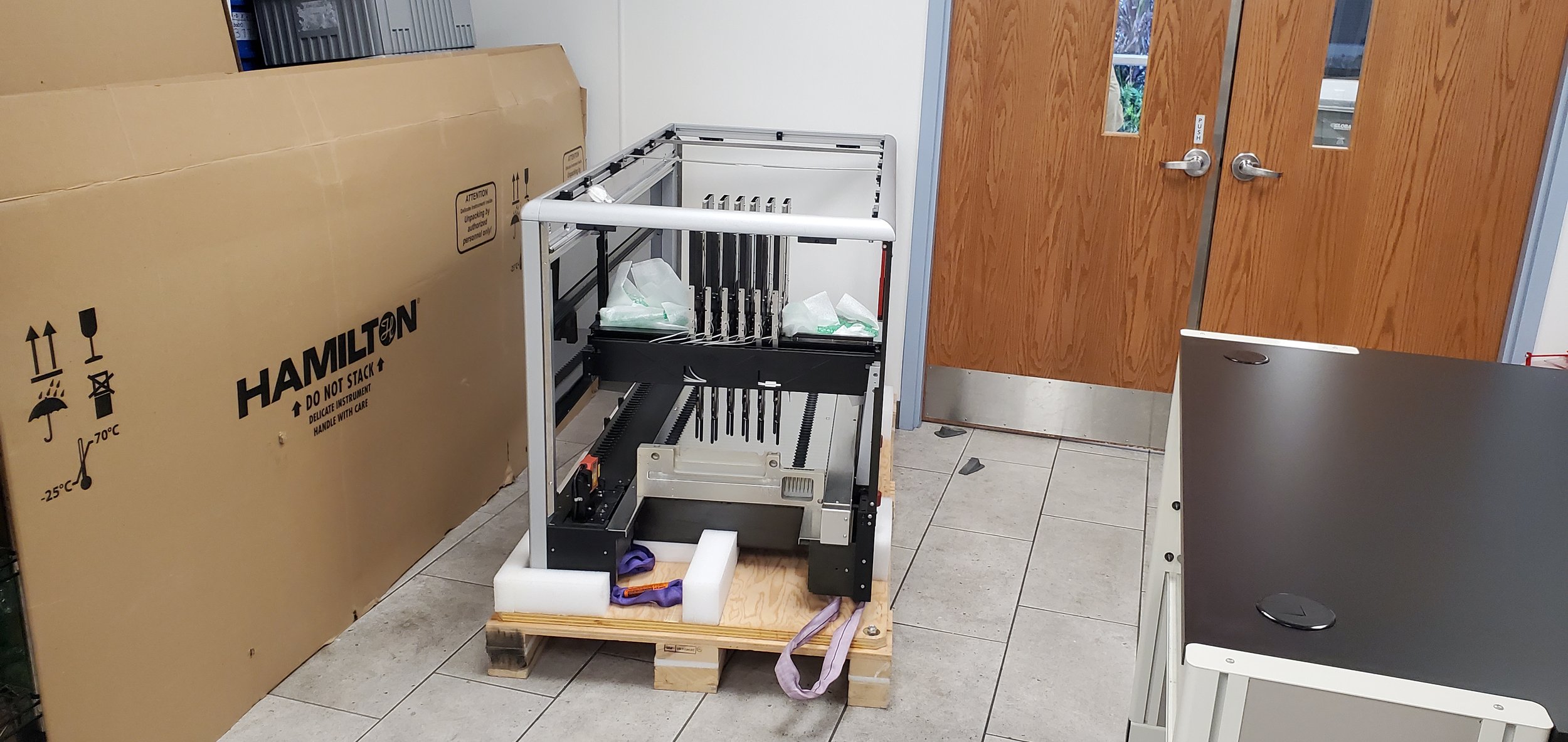 Professional Handymen Service, An empty lab or office room with a partially assembled 3D printer on a wooden pallet, a large cardboard box labeled Hamilton, and two wooden doors with windows. The room has tiled flooring and white walls.