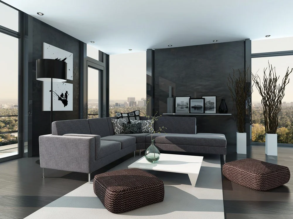 Modern living room with gray sectional sofa, black and white wall art, a white coffee table, two woven poufs, large windows showing city skyline, and decorative vases.