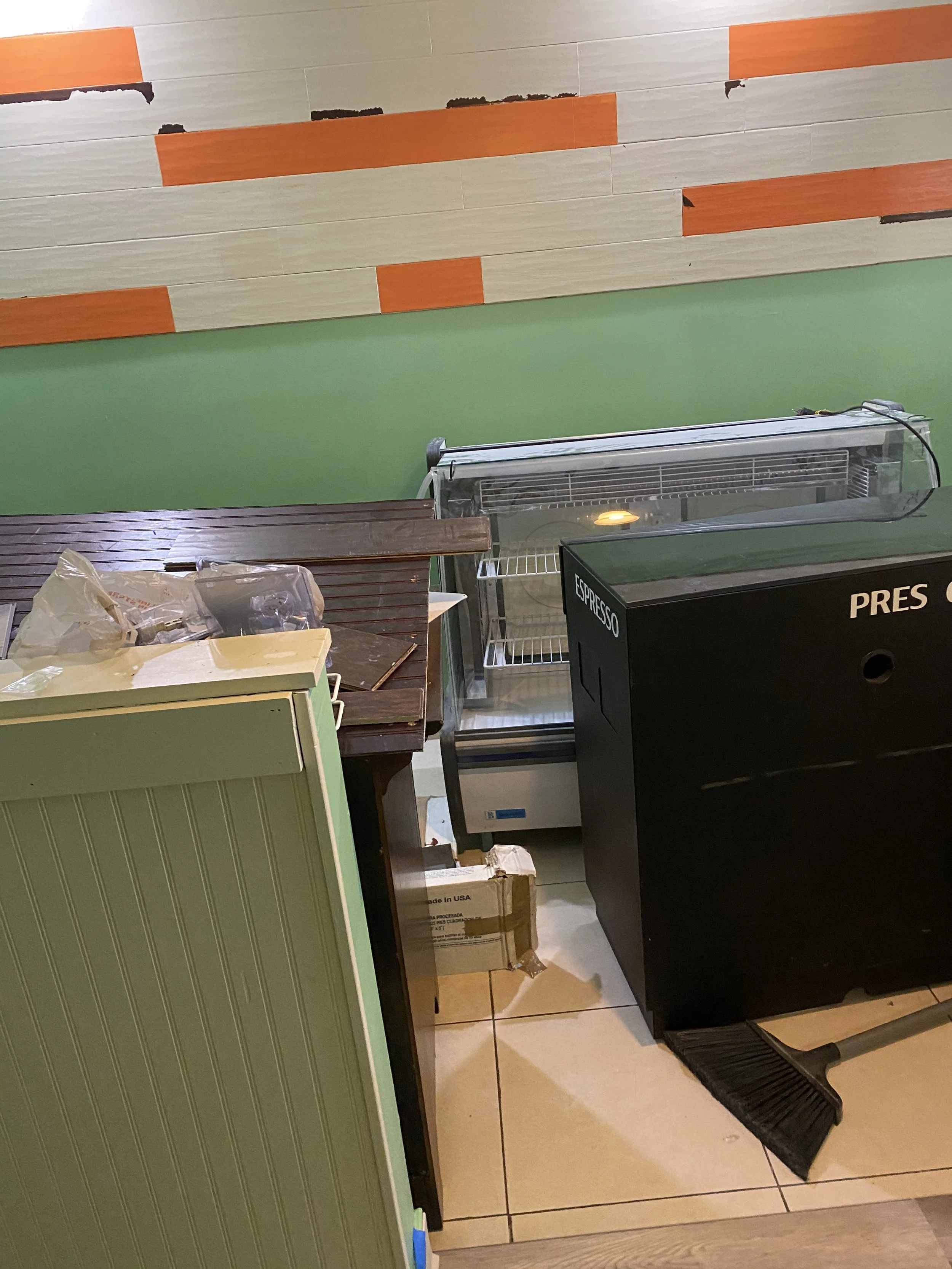 Part of a room with a green wall, a wooden counter, and a black espresso machine. There are boxes, paper bags, and a glass display case or refrigerator, some items are on the floor and the counter. The floor is tiled.