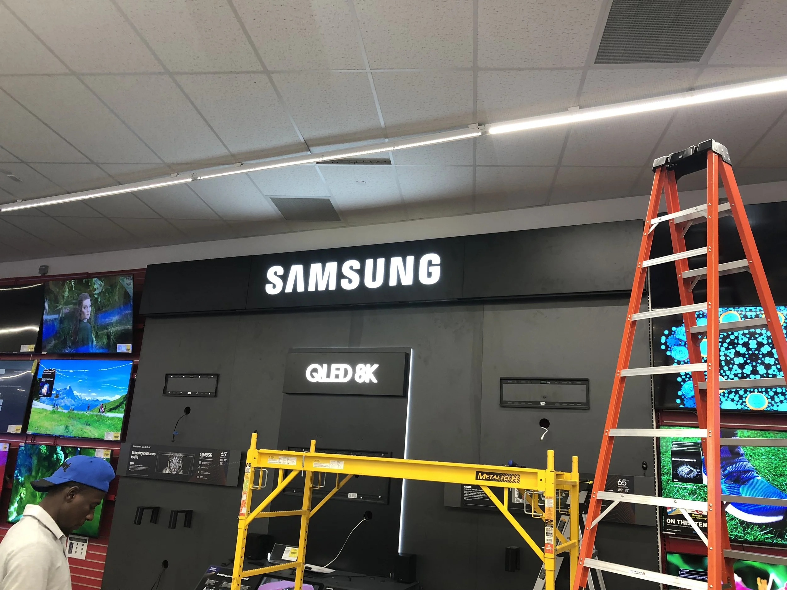 Interior of an electronics store with a Samsung sign, a ladder, a scaffold, and multiple TV displays on the wall.