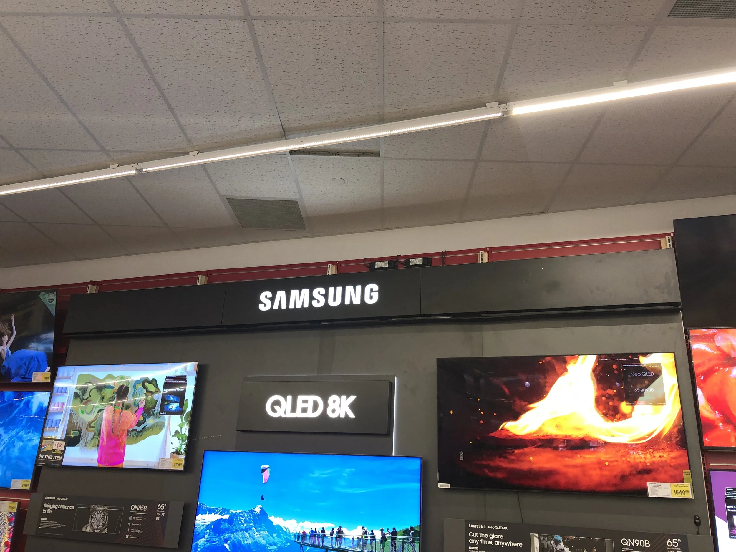 Inside an electronics store showcasing Samsung televisions, including models QLED 8K and Neo QLED 4K, with colorful screens and promotional signage.