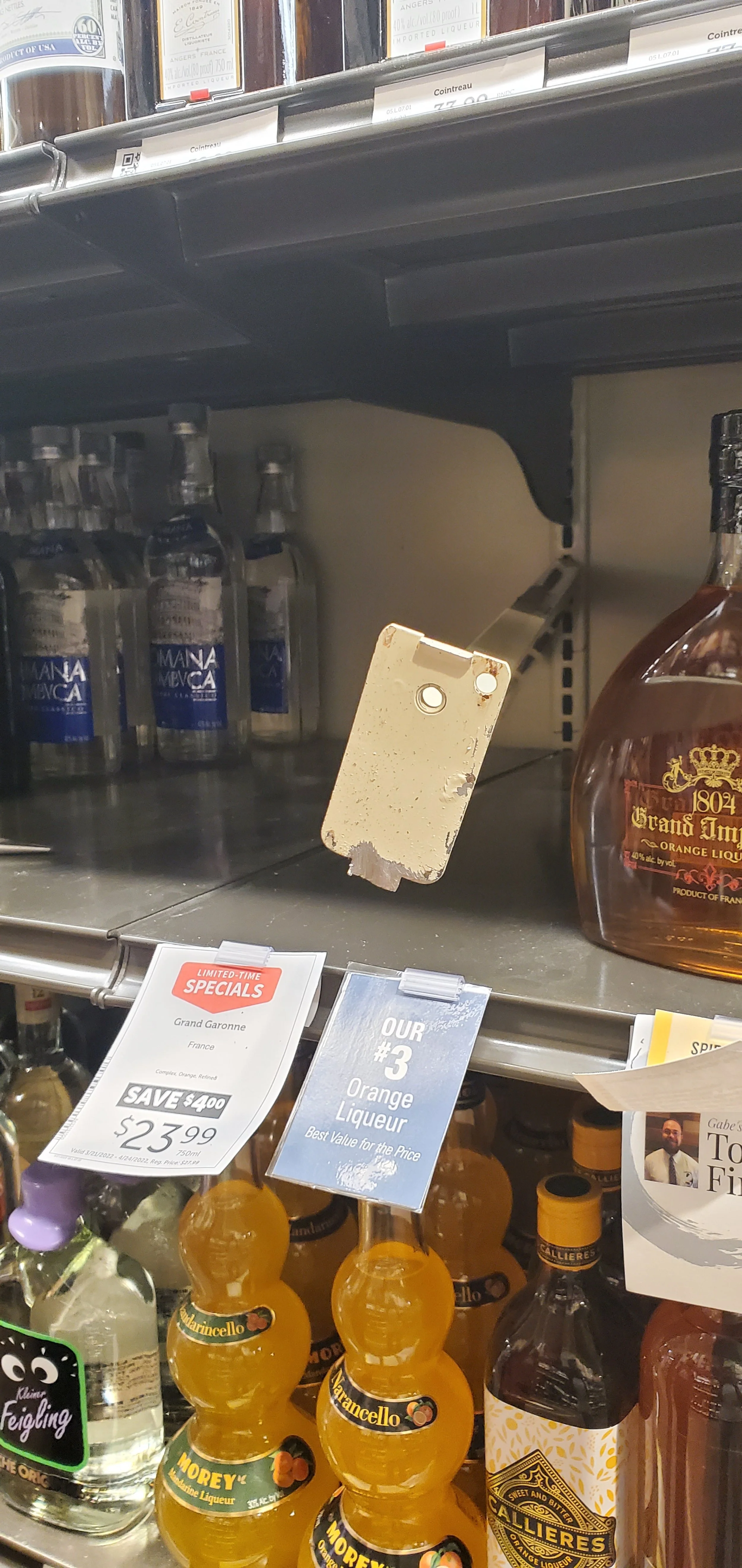 Empty retail shelf with some bottles of alcohol, including a bottle of Grand Marnier and Callieres orange liqueur, with price tags and promotional signs.