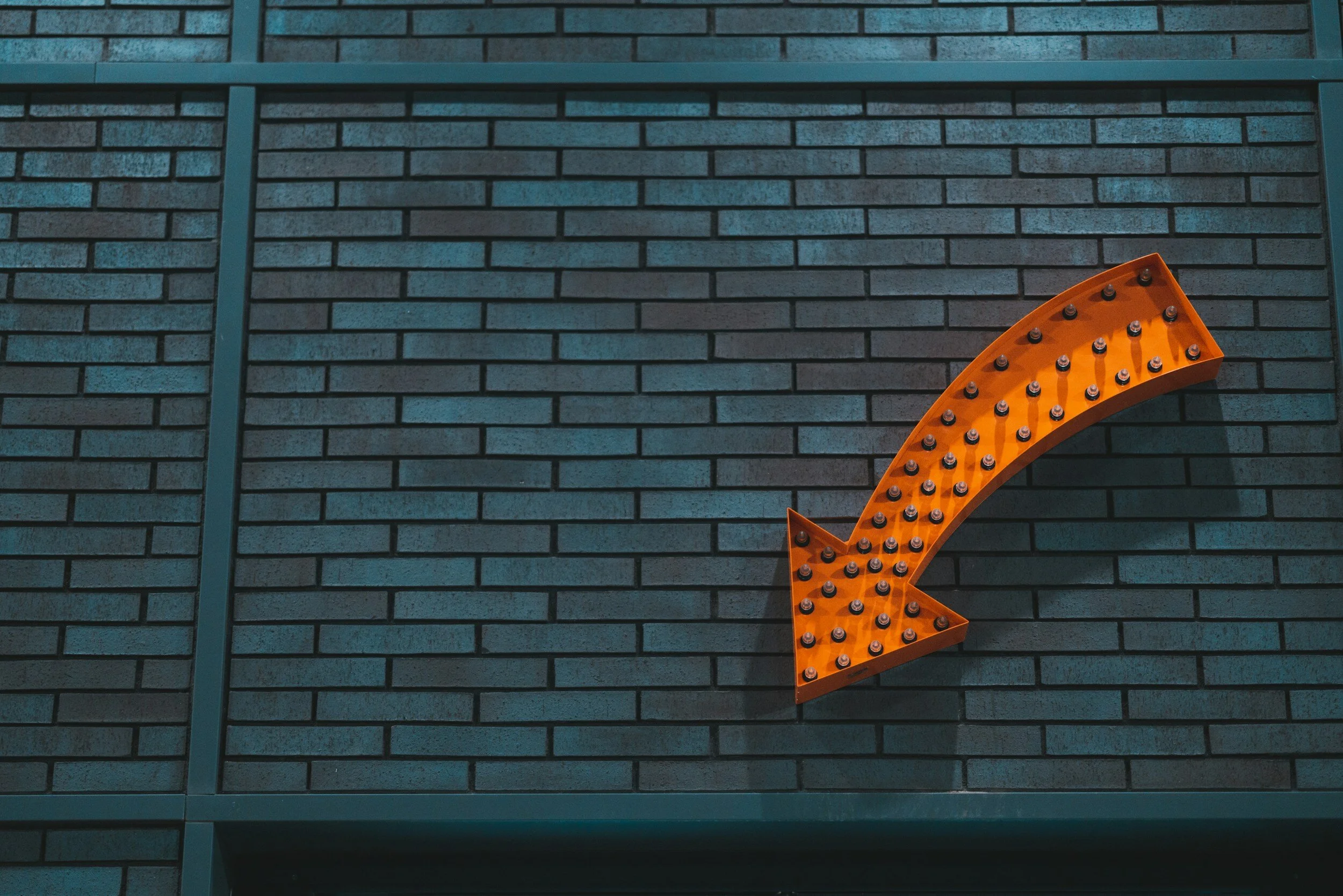 Orange arrow sign with bulbs pointing right, mounted on a dark brick wall.