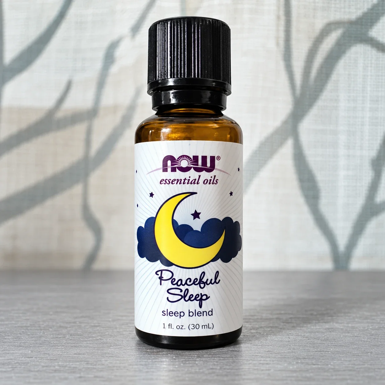 NOW Peaceful Sleep essential oil blend bottle against a softly lit curtain