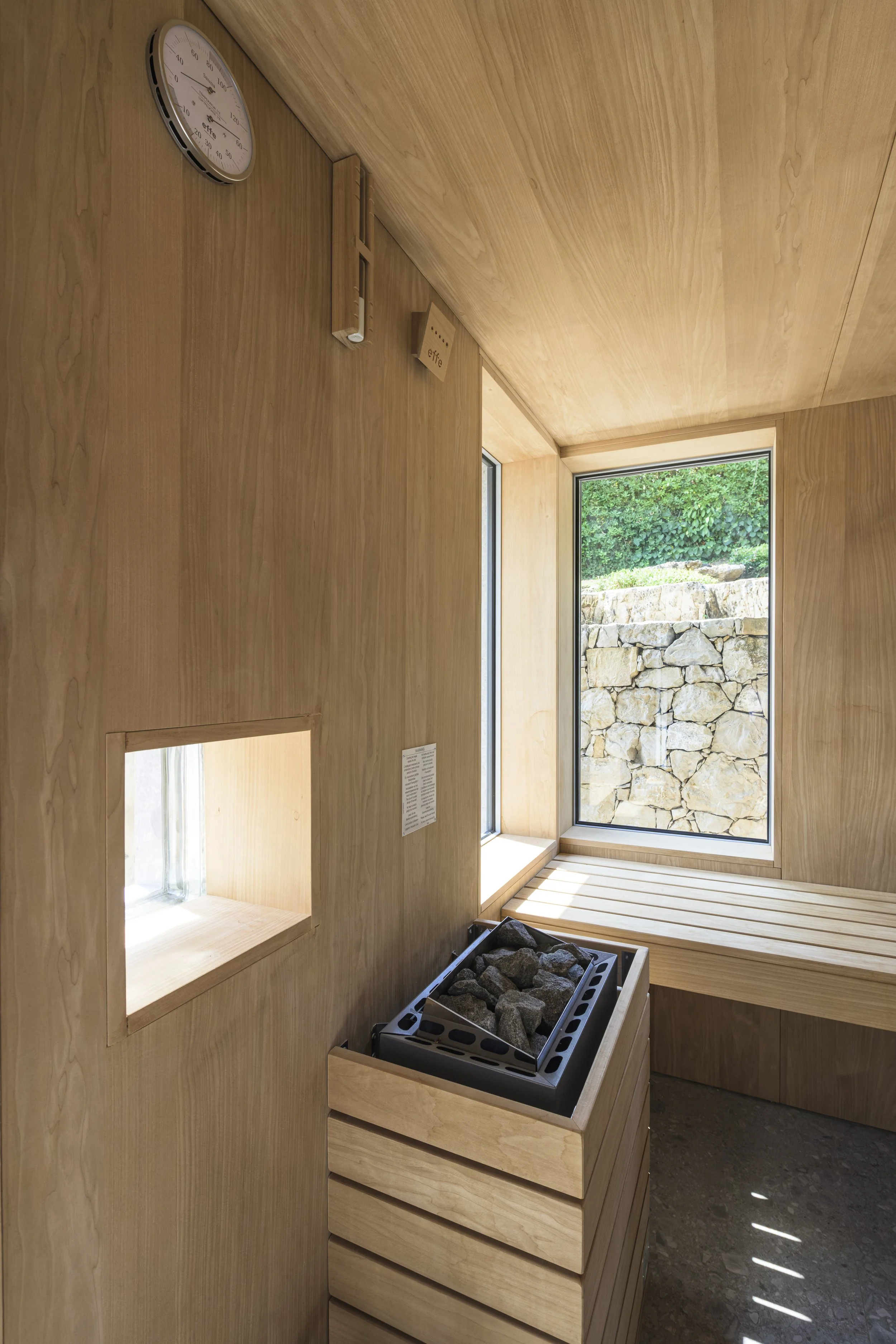 Intérieur du sauna : boiserie claire, et une grande baie qui cadre le mur en moellons conservé comme un tableau.