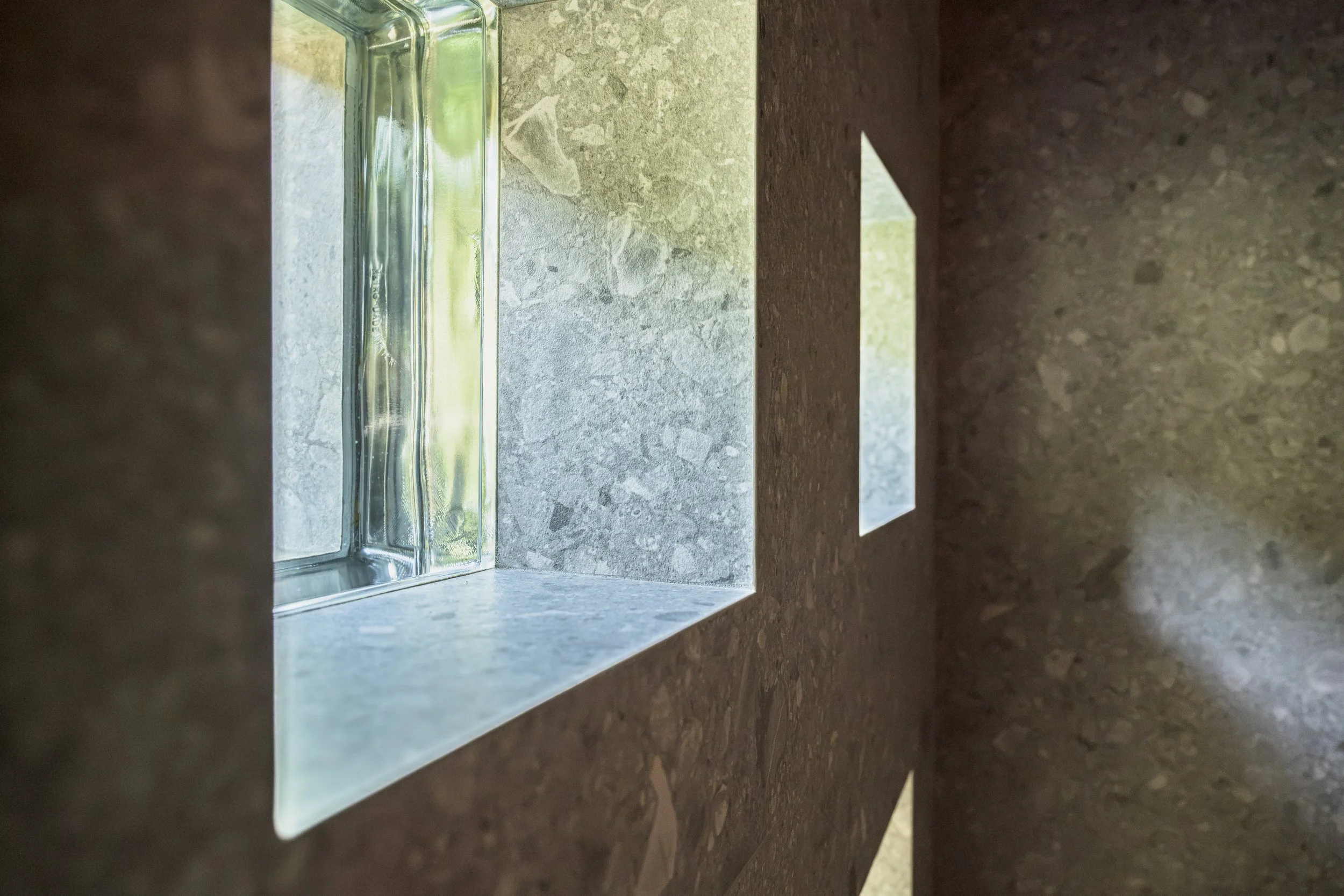 Pavés de verre vus depuis le hammam. Le revêtement se retourne dans l'épaisseur du mur et habille l'ébrasement sur ses quatre faces, sans profilé ni rupture : la même matière court du sol aux parois, de l'intérieur à l'extérieur. Les arêtes vives en 