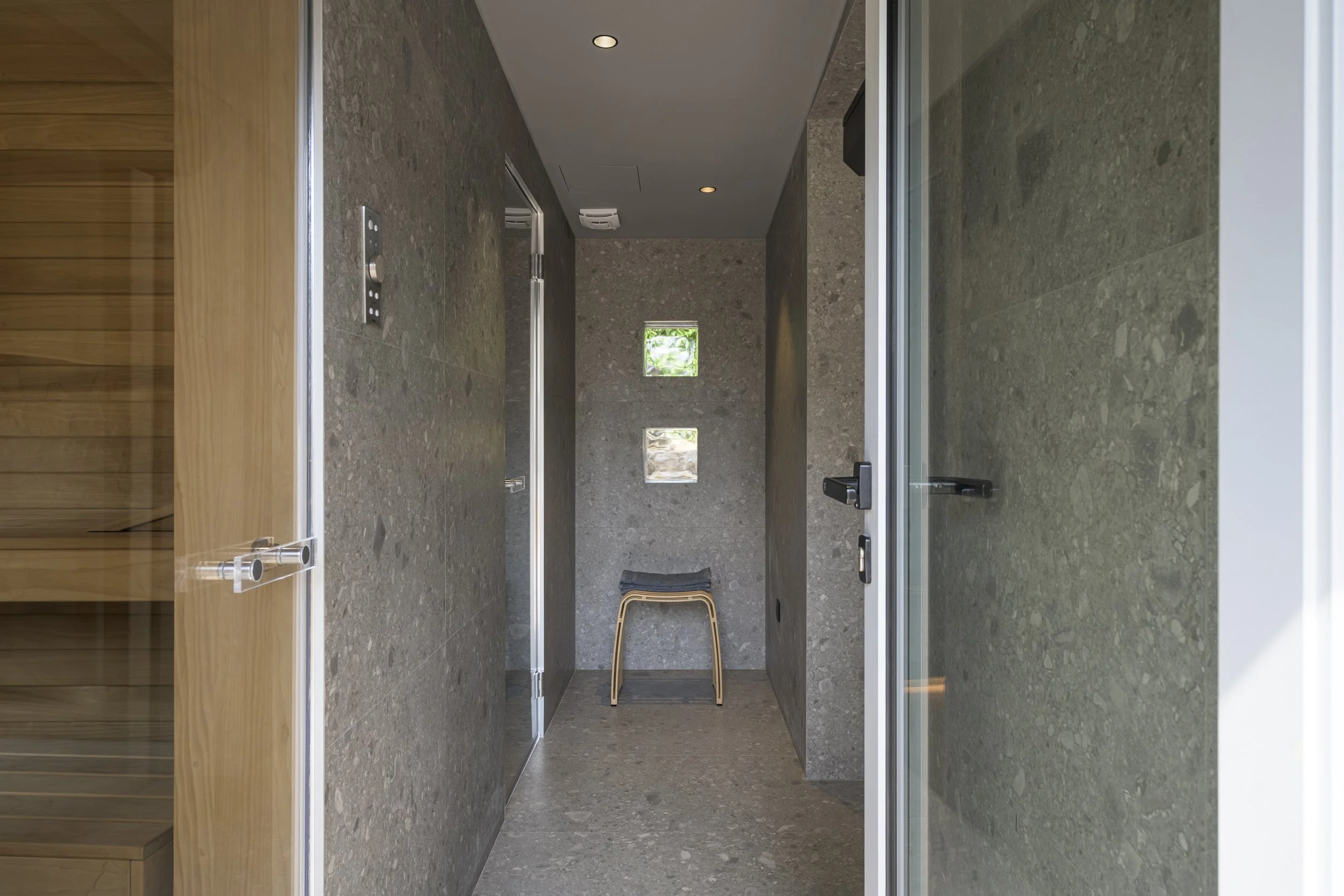 Vue intérieure depuis l'entrée : le sauna en bois à gauche, le hammam derrière la porte vitrée, et au fond les pavés de verre qui laissent entrer la lumière du jardin. Sol et murs dans le même revêtement minéral qu'à l'extérieur.