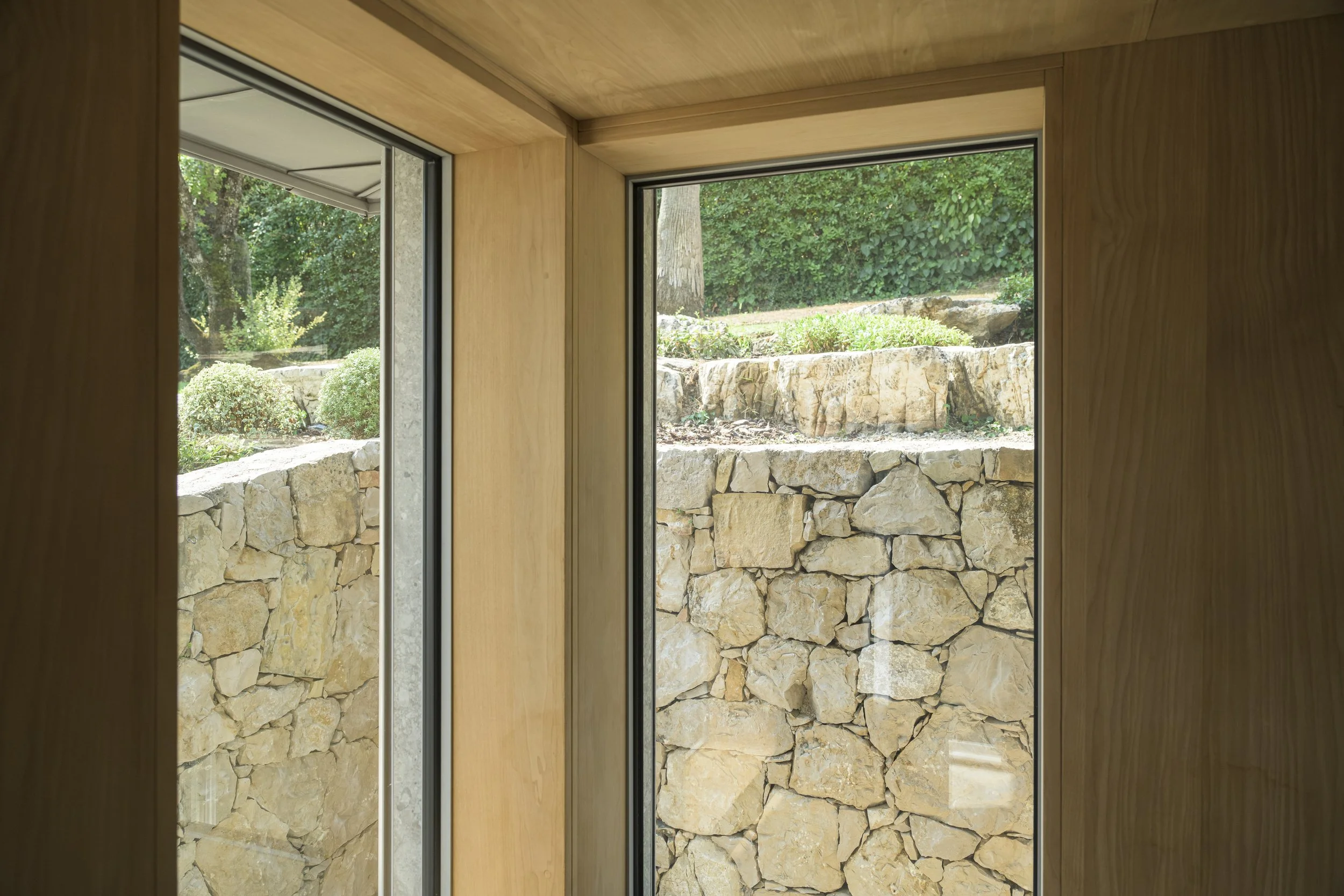 Angle vitré du sauna : deux baies encadrées de bois révèlent l'étagement du site, du mur en moellons aux terrasses du jardin.