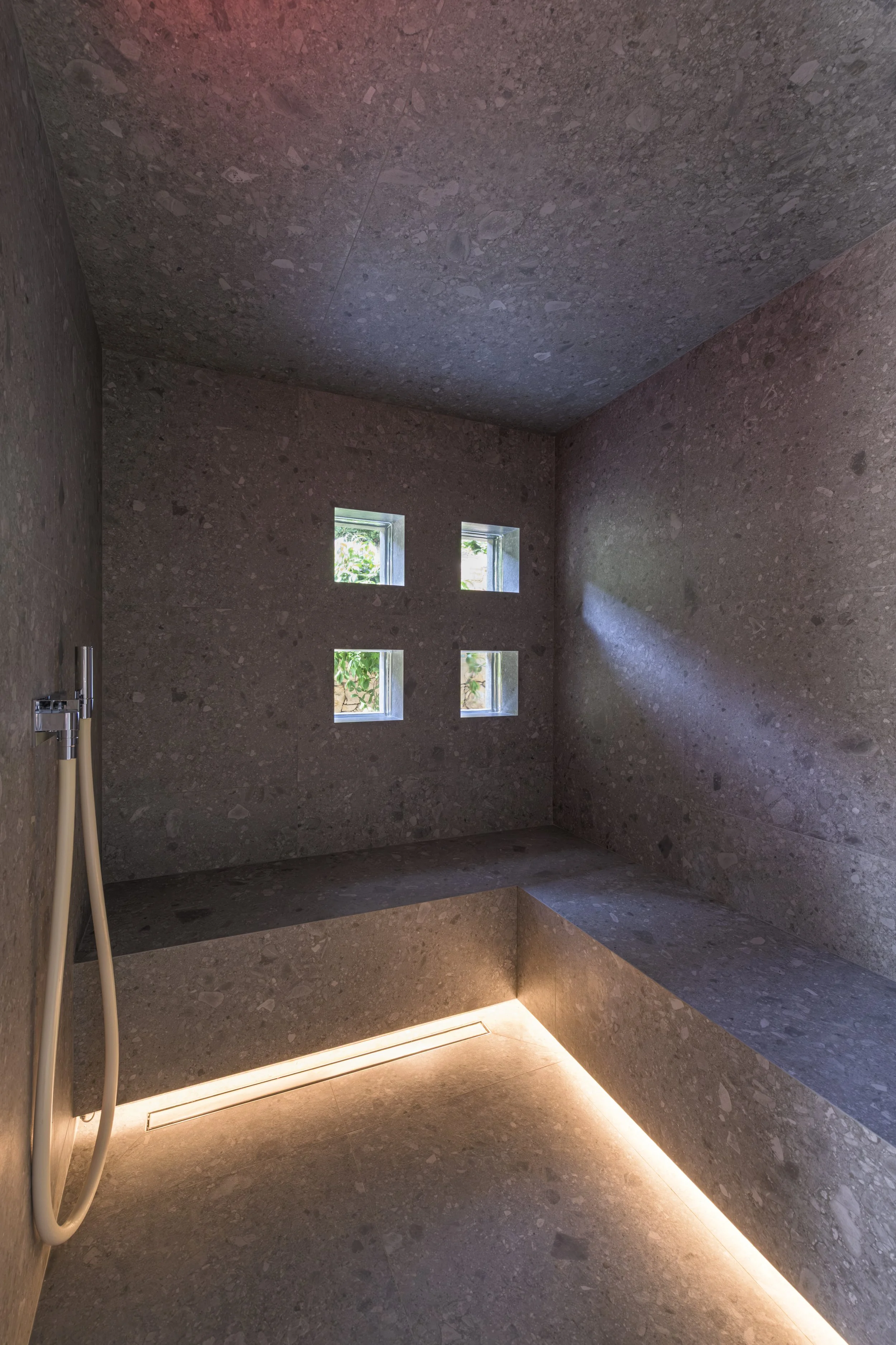 Intérieur du hammam : banquette dans le même revêtement minéral, éclairée à sa base par une ligne de lumière chaude, tandis que les pavés de verre laissent entrer la lumière du jardin.