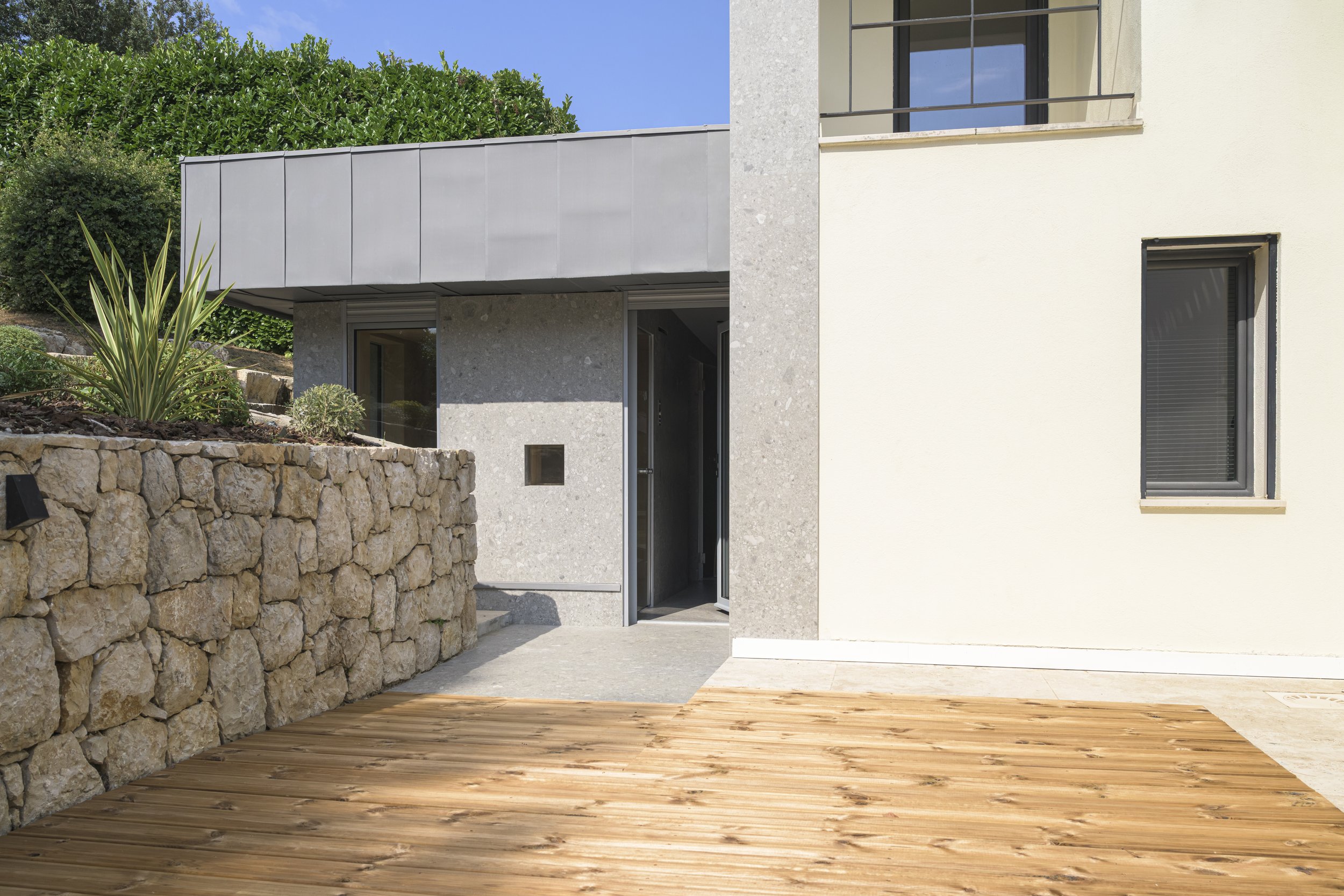 Vue d'ensemble depuis la terrasse : l'extension s'insère entre le mur en moellons conservé et la villa existante, reliée à celle-ci par une bande du même revêtement minéral.