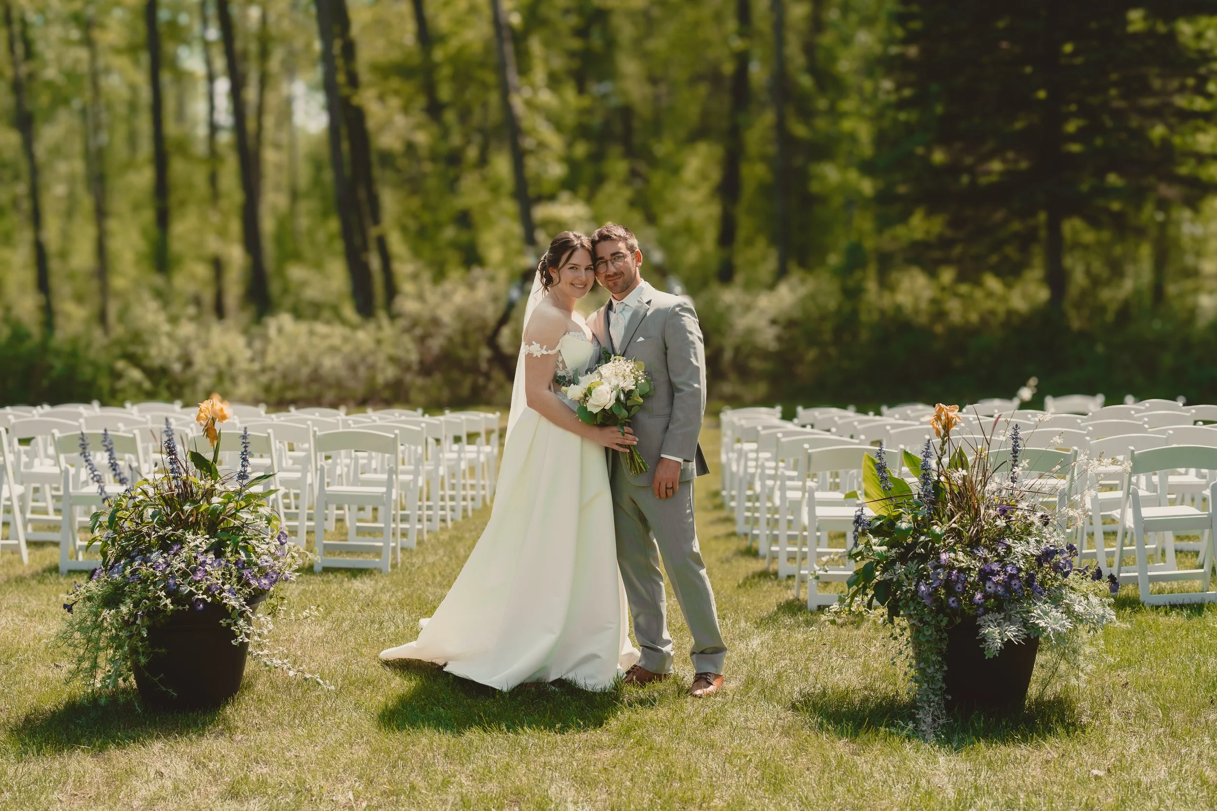 A bride and groom standing together outdoors on a grassy area, surrounded by wedding chairs and flower arrangements, with a forested background on a sunny day.