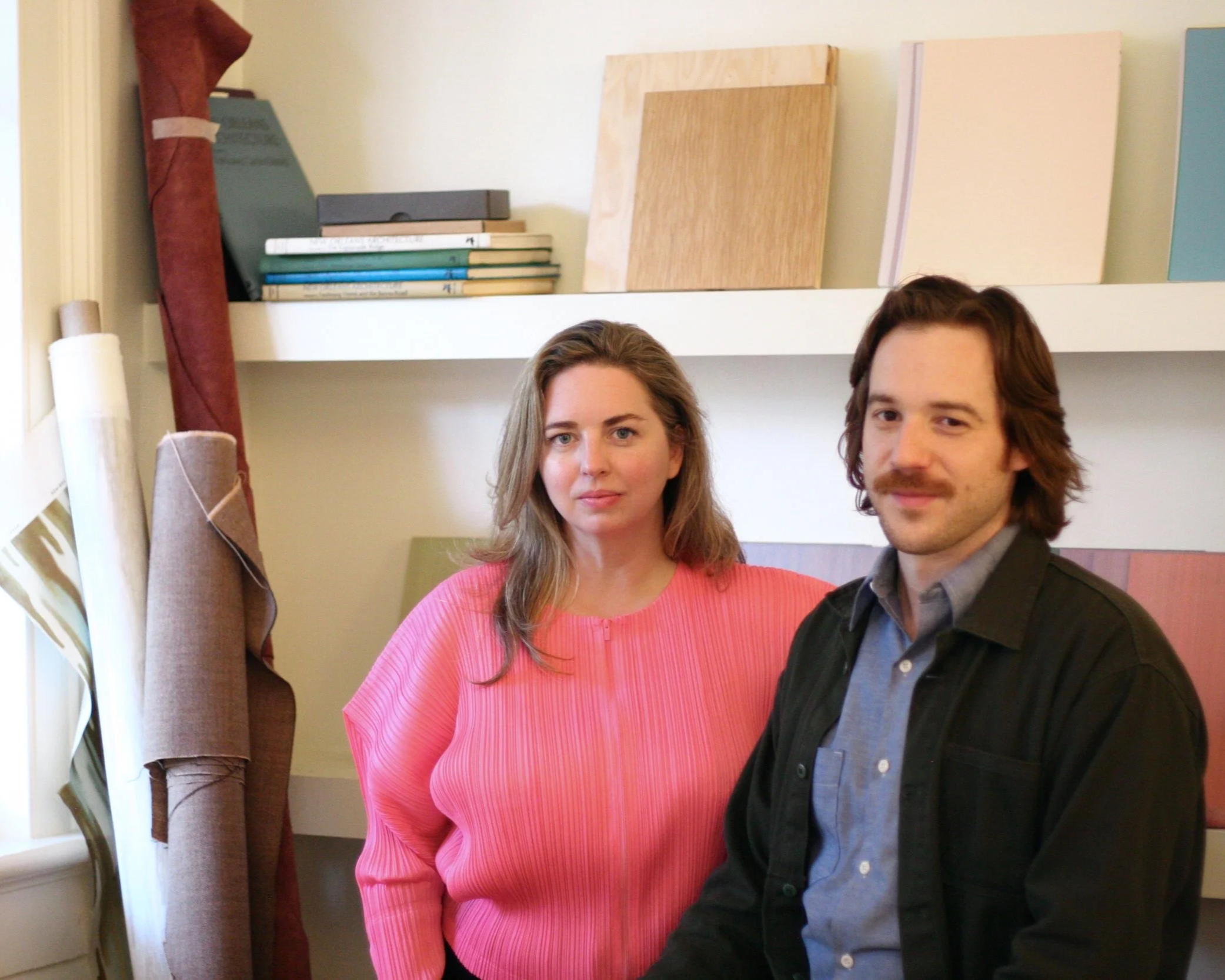 Two young adults standing side by side indoors. The woman is wearing a pink ribbed sweater and has shoulder-length light brown hair. The man has wavy brown hair, a mustache, and is wearing a dark jacket over a gray shirt. They are standing in front of a wall with colorful rectangular panels and shelves, with some fabric and rolls of paper or fabric on the left side.