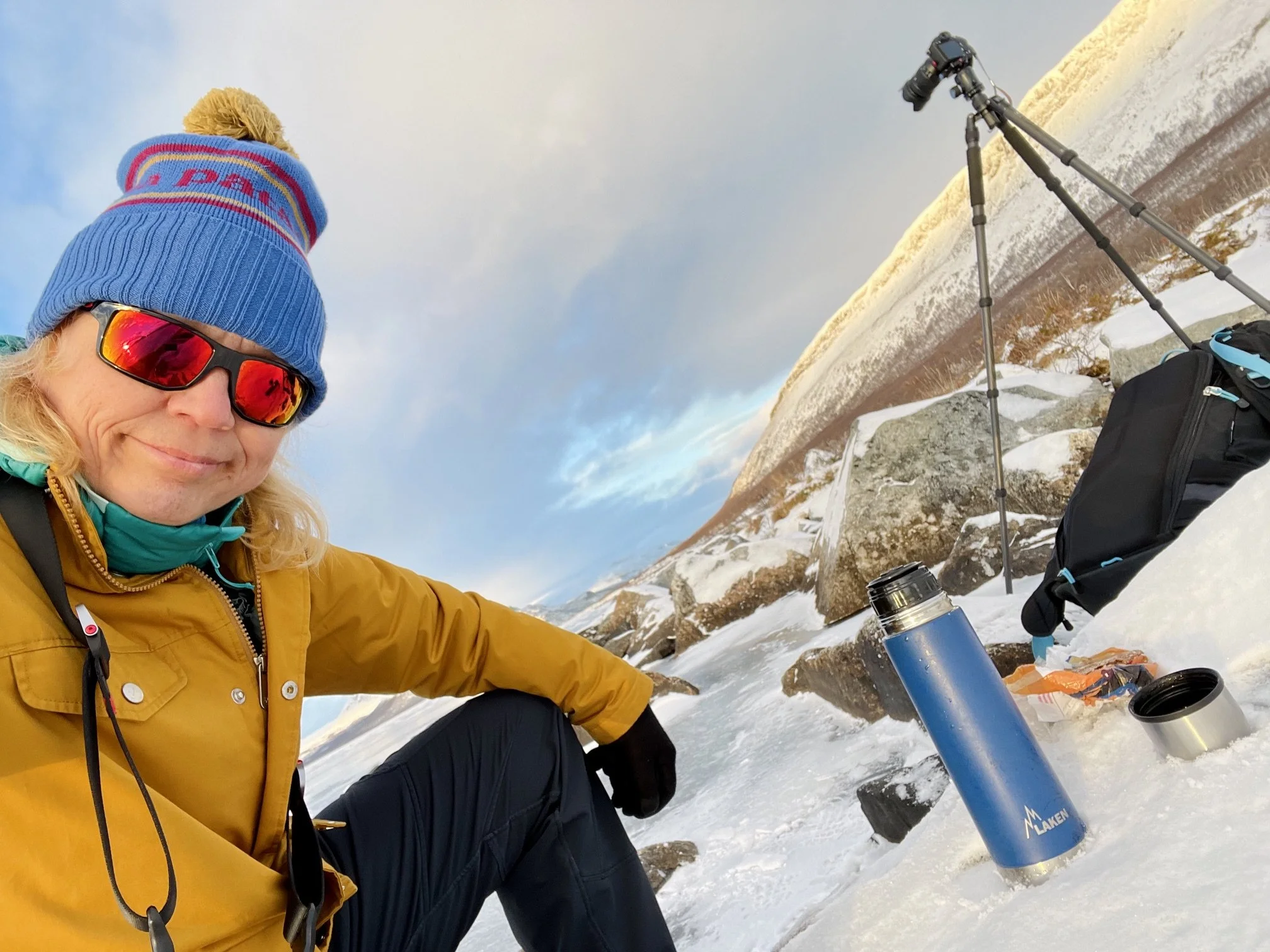 Selfie during a snowy morning break in Lapland, with a thermos and sandwich in the foreground and a camera and tripod standing in the snow behind.