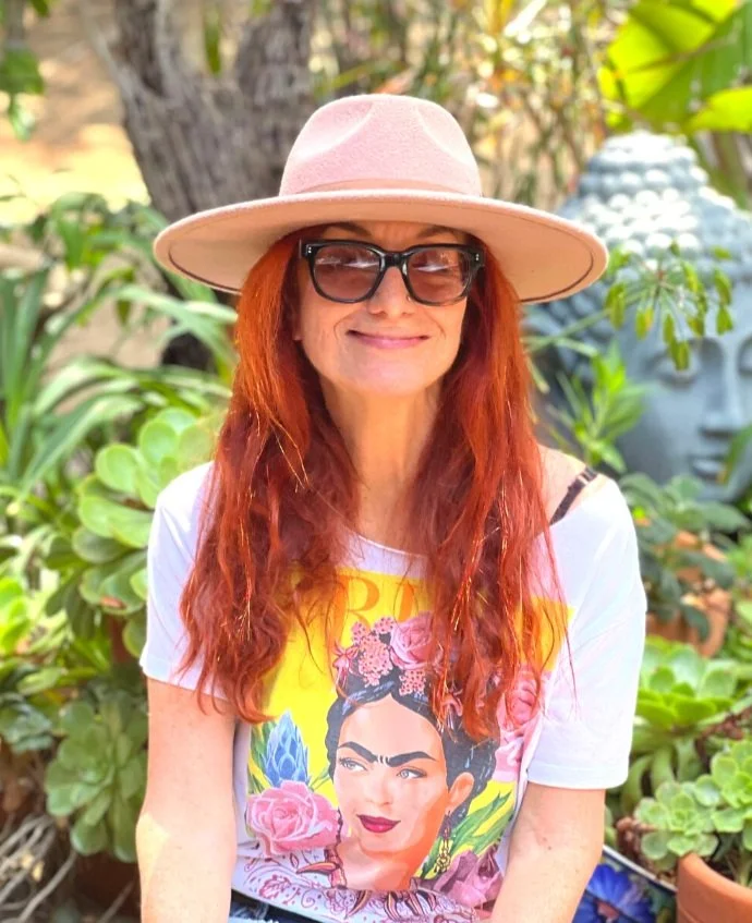 Woman with long red hair wearing a pink wide-brimmed hat, black sunglasses, a graphic Frida Kahlo T-shirt, and a black strap, standing outdoors among green plants.