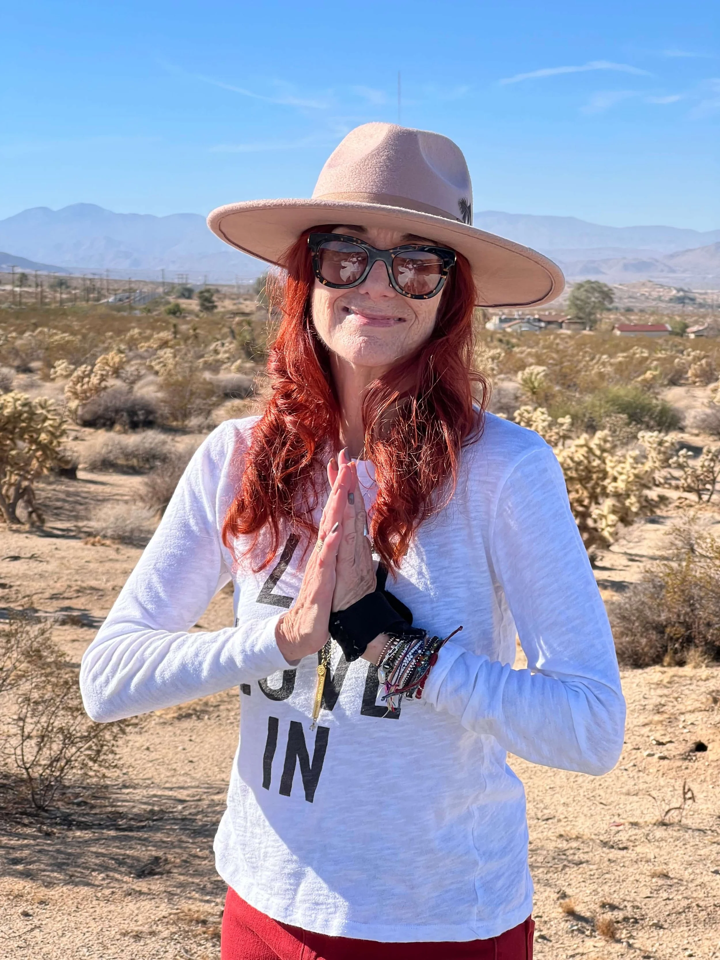 A woman with red hair wearing a wide-brimmed hat, sunglasses, and a white long sleeve shirt stands in a desert landscape with mountains and scrub bushes in the background. She holds her hands together in a prayer position.