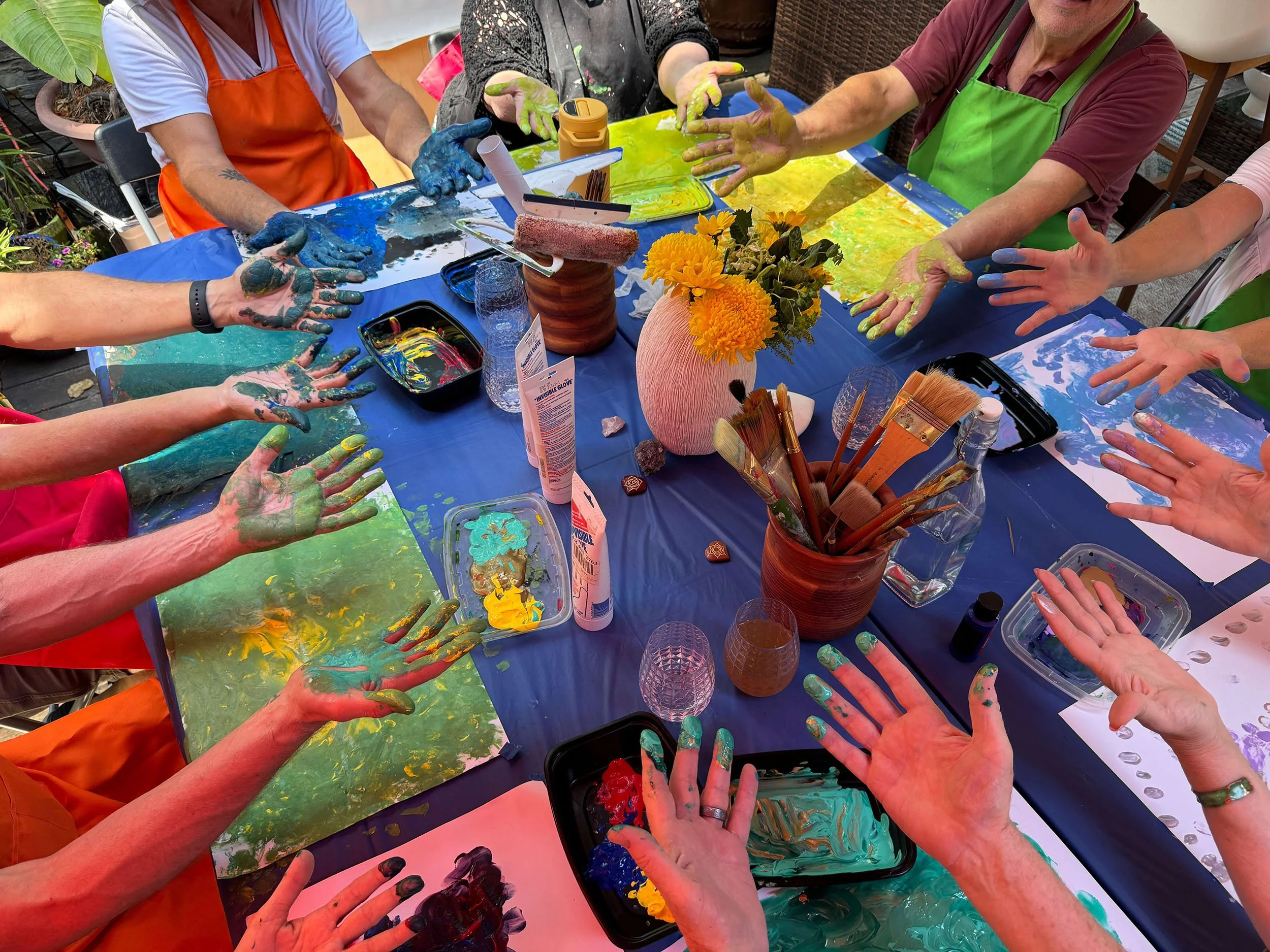 People with paint-covered hands participating in a painting activity around a table with art supplies, including paintbrushes, paint palettes, and paper with colorful paint splashes, during a fun outdoor gathering.