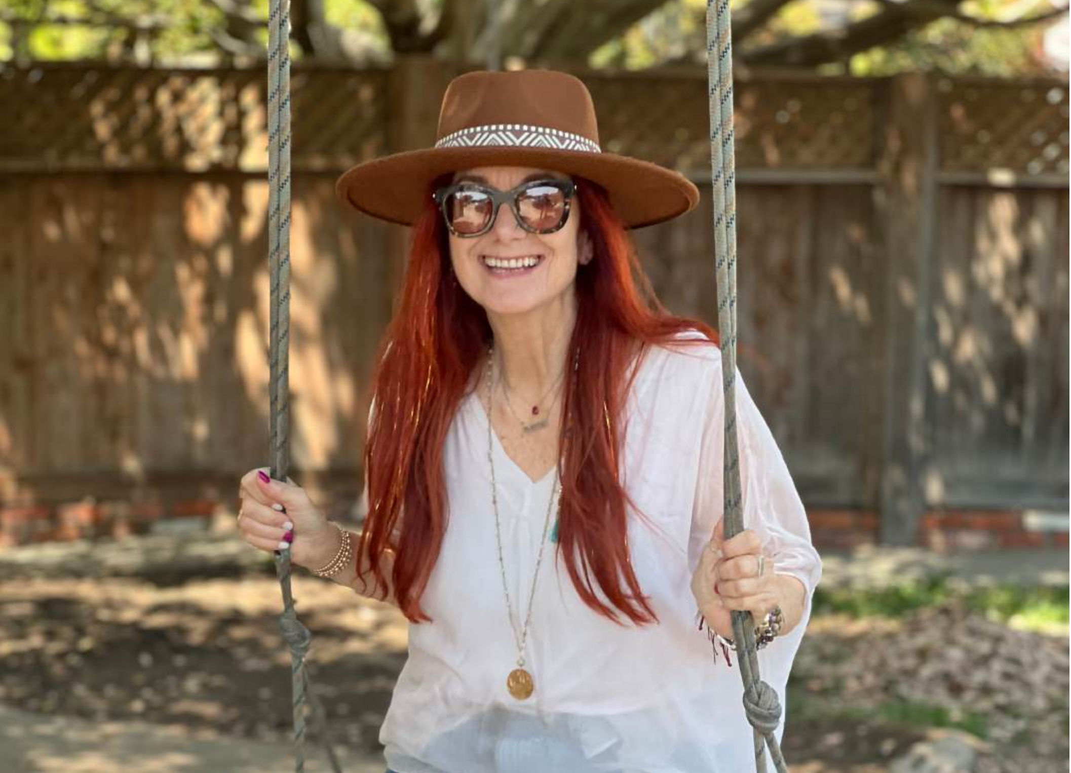 Jessie Marcus wearing a wide-brimmed brown hat, sunglasses, a white blouse, and jewelry, holding onto swing ropes in backyard with wooden fence and trees in background.