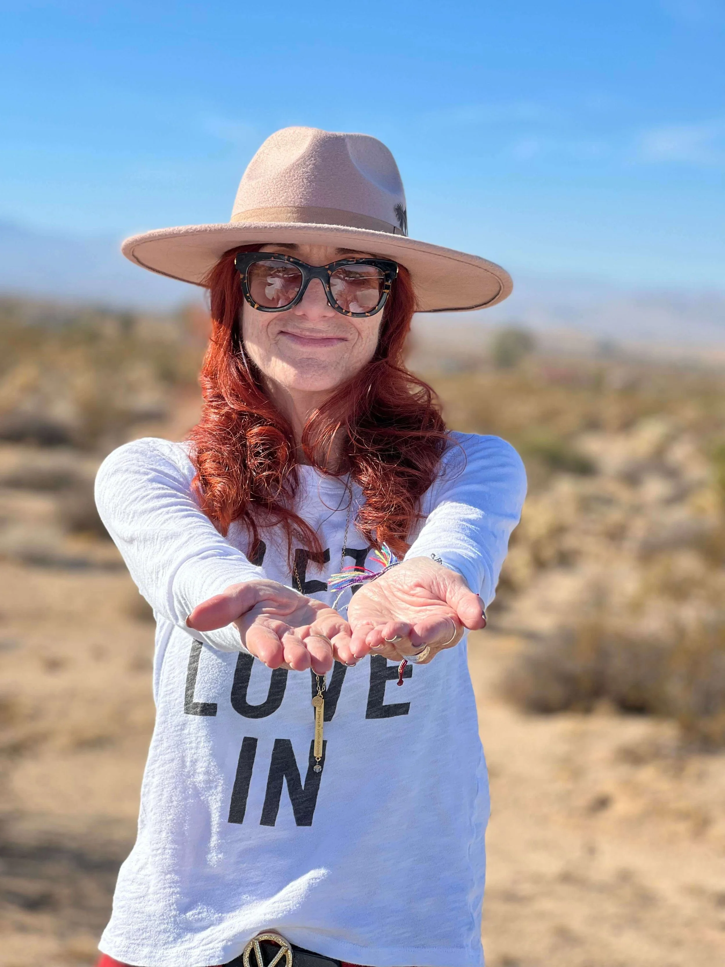 Jessie Marcus wearing a beige wide-brim hat, black sunglasses, a white long-sleeve shirt with the text 'LOVE IN', and standing outdoors in a desert landscape with a blue sky.