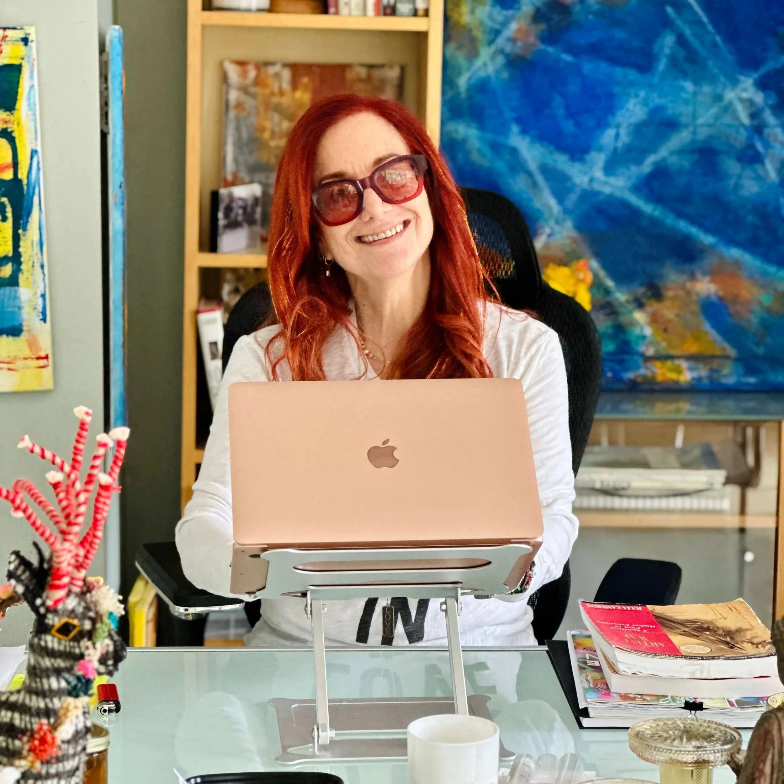 Jessie Marcus wearing glasses, smiling while sitting at a desk with a pink MacBook. The background features colorful abstract art and a bookshelf.