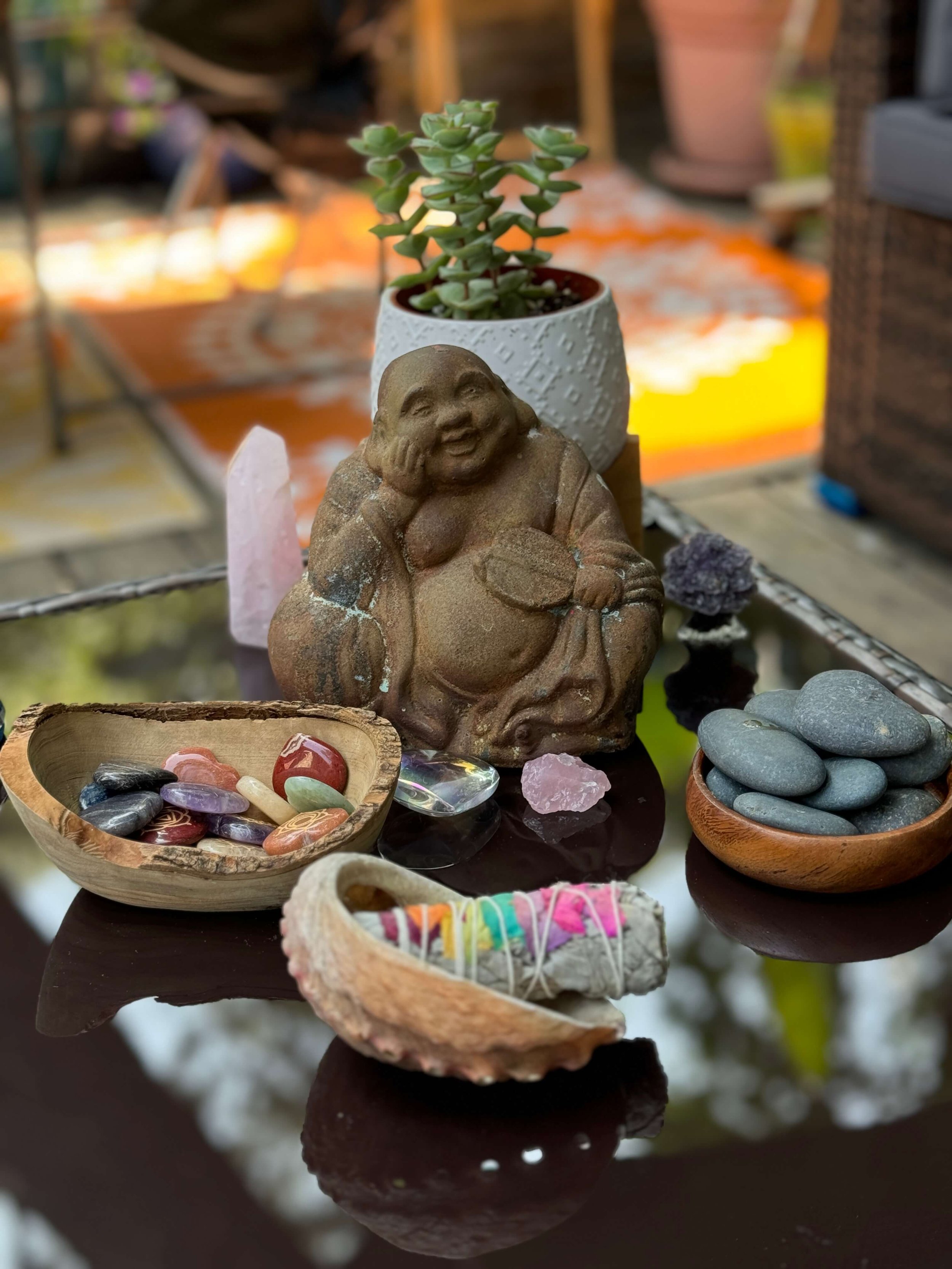 A collection of decorative items including a laughing Buddha statue, a white potted succulent, several polished stones in bowls, a small colorful wrapped item, and crystals on a reflective black surface.