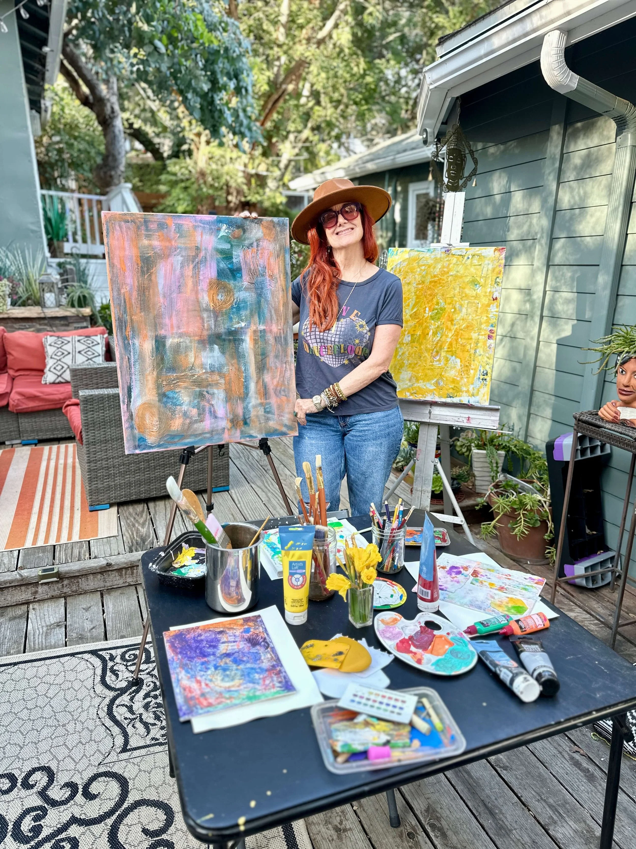 Jessie Marcus wearing a brown hat, sunglasses, and a graphic T-shirt standing outdoors at an art studio, smiling, with paintings and art supplies on a table and easels behind her.