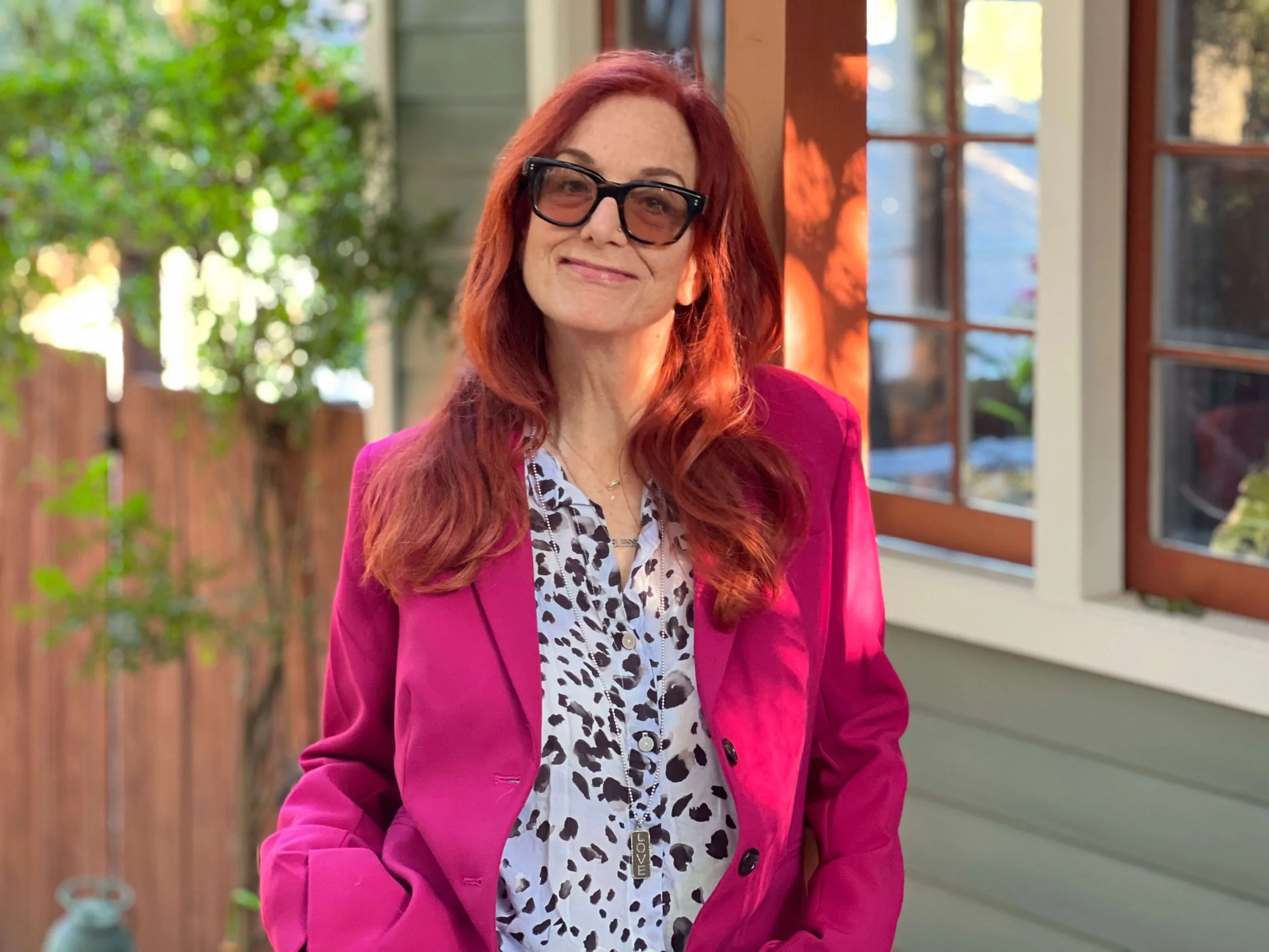 Jessie Marcus wearing a bright pink blazer over a white blouse with black spots, is standing outside near a window with a wooden frame. The background includes a wooden fence and greenery, with sunlight illuminating her face.