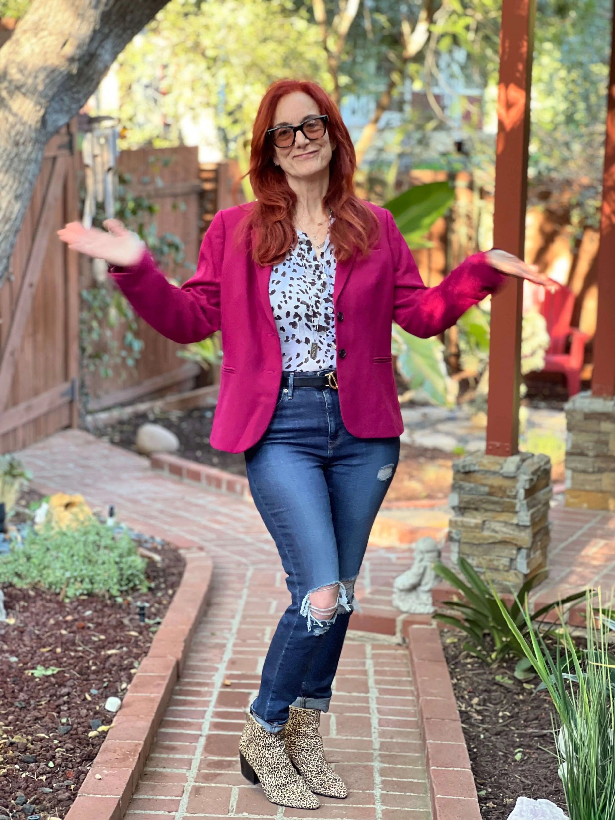 Jessie Marcus standing on a brick pathway in a garden with trees and plants, wearing a magenta blazer, animal print blouse, ripped jeans, and leopard print ankle boots, with arms raised and palms facing up.