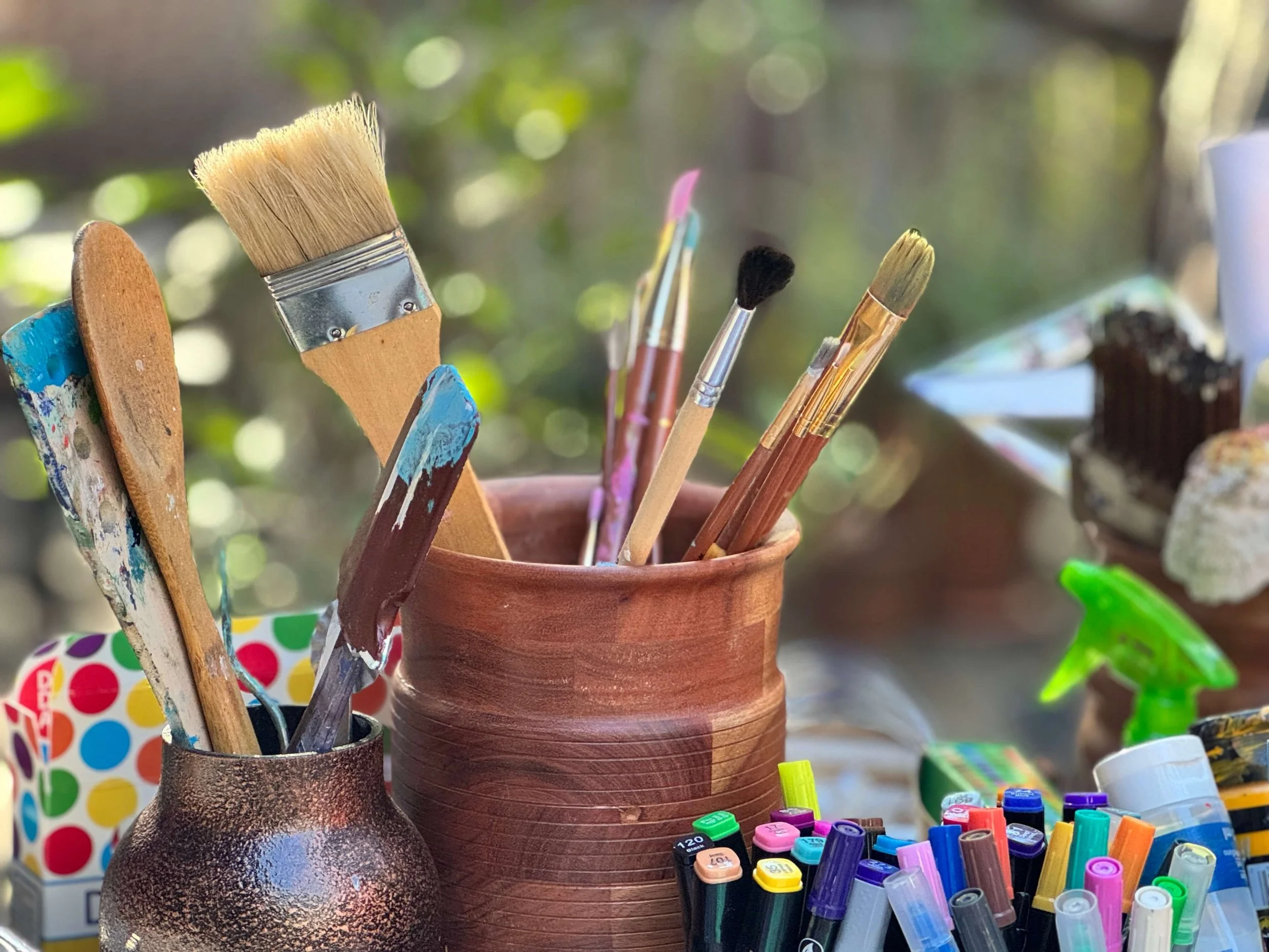 An assortment of paintbrushes and markers in containers, with a spray bottle and other art supplies on a table outdoors.