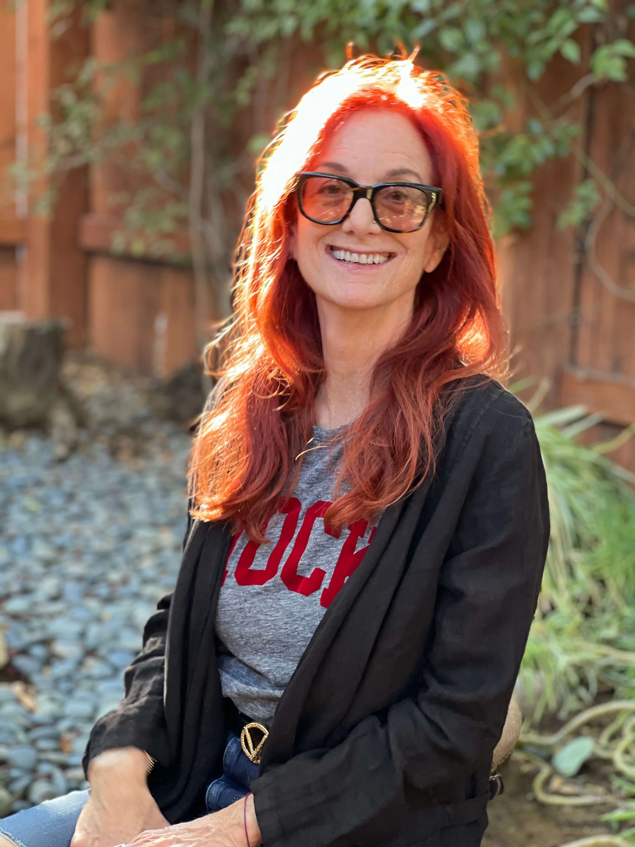 Jessie Marcus wearing glasses smiling outdoors during golden hour, sitting in front of a wooden fence with green foliage, wearing a gray t-shirt and a black jacket.