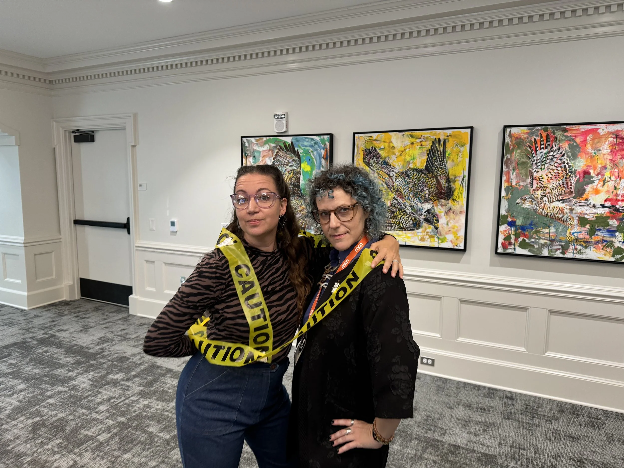 Two white women wearing safety goggles look mysteriously at the camera while wrapped in a strand of yellow CAUTION tape.