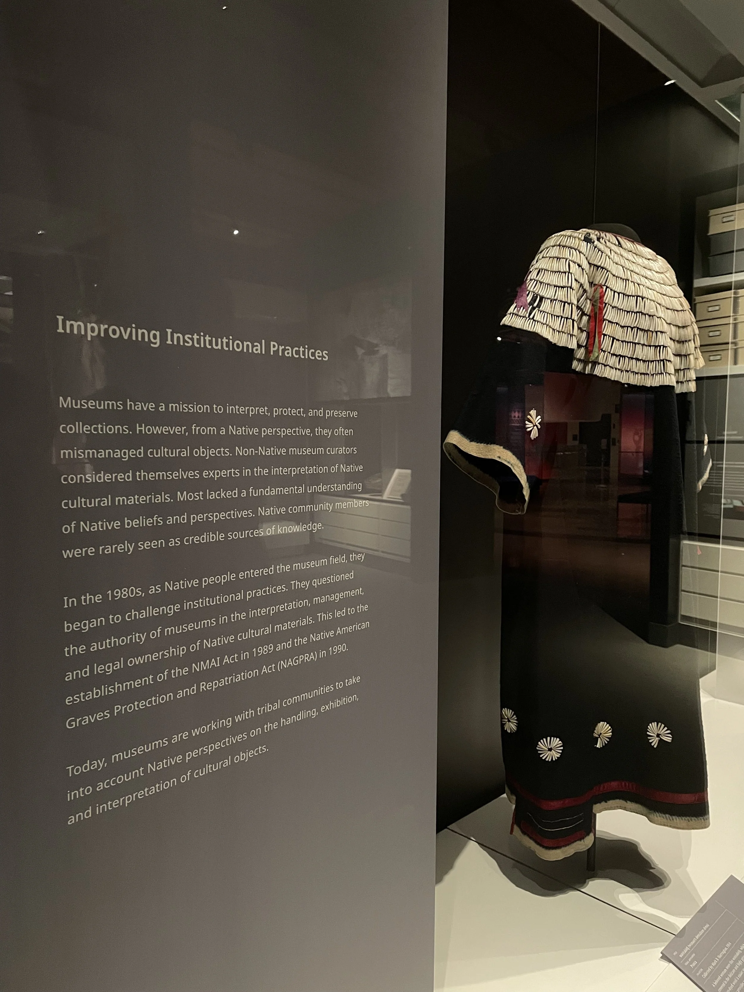 A photo of a museum case with an embellished dress next to a label titled "Improving Institutional Practices" that explains some of the history of museums marginalizing Native people.