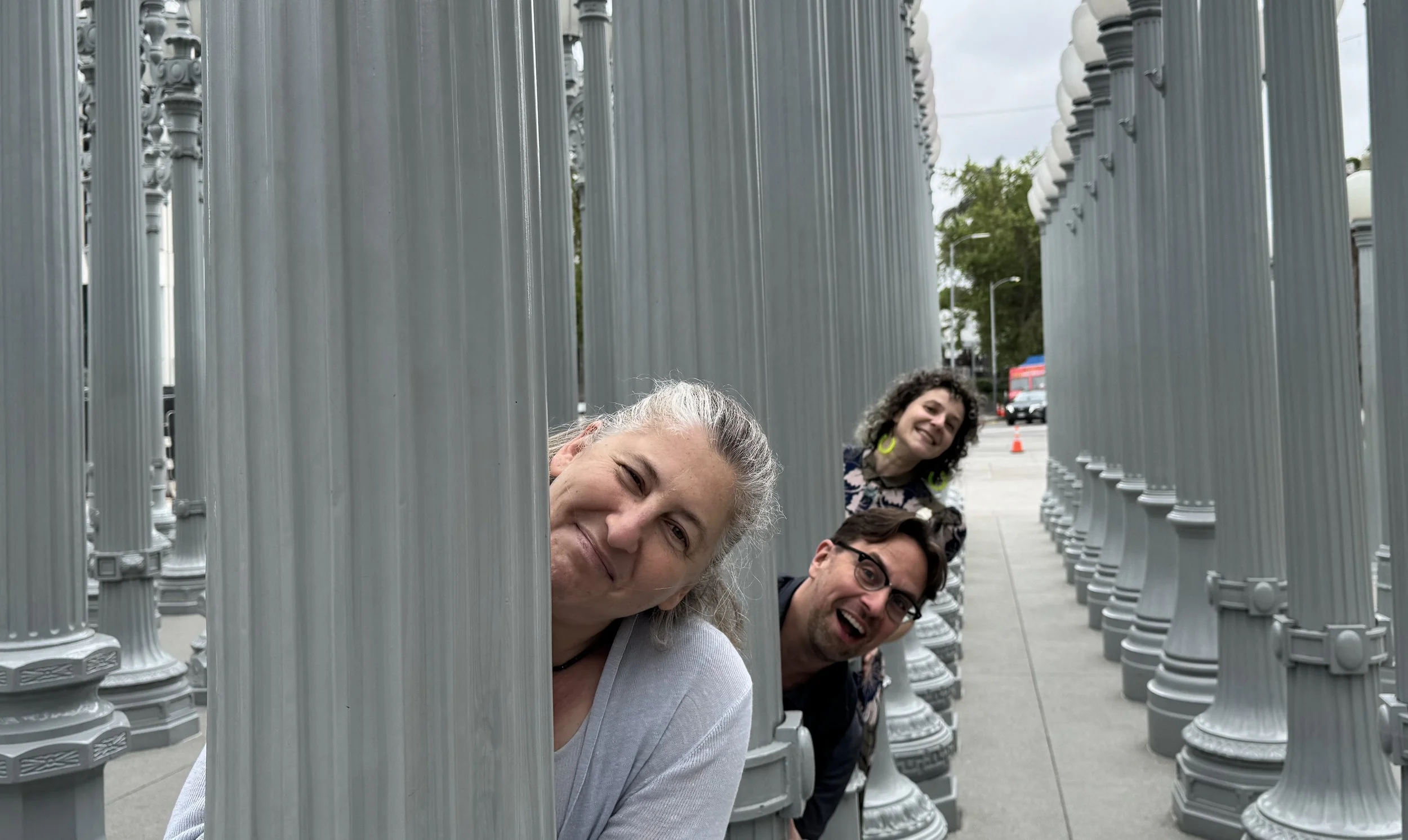 Three smiling faces peek out between a forest of gray lamp posts on a sidewalk.