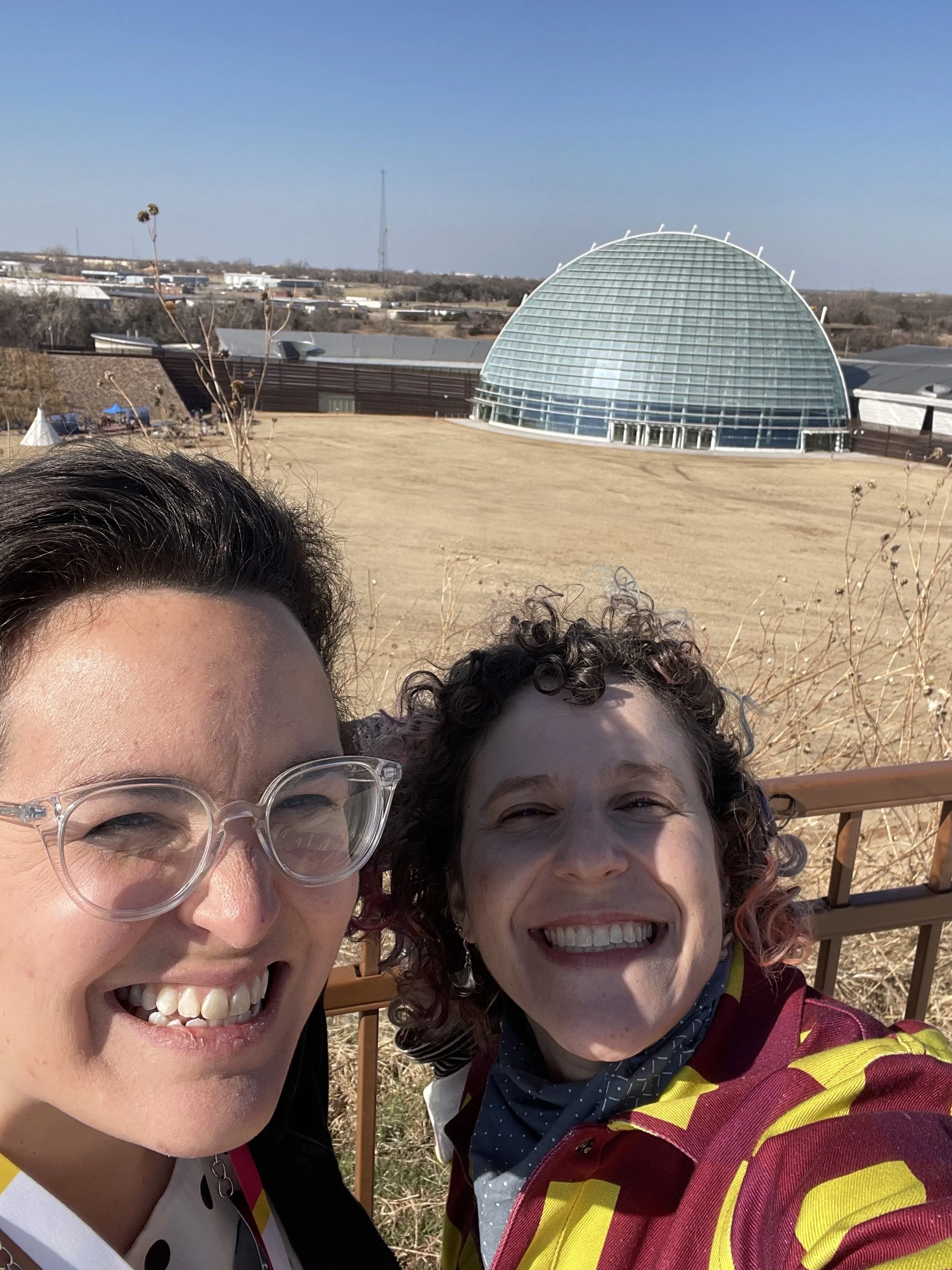 A selfie of two squinting, smiling faces of pale-skinned ladies (me and Adrienne) with the domed, paned glass structure of the First Americans Museum rising over an expanse of plains in the background.