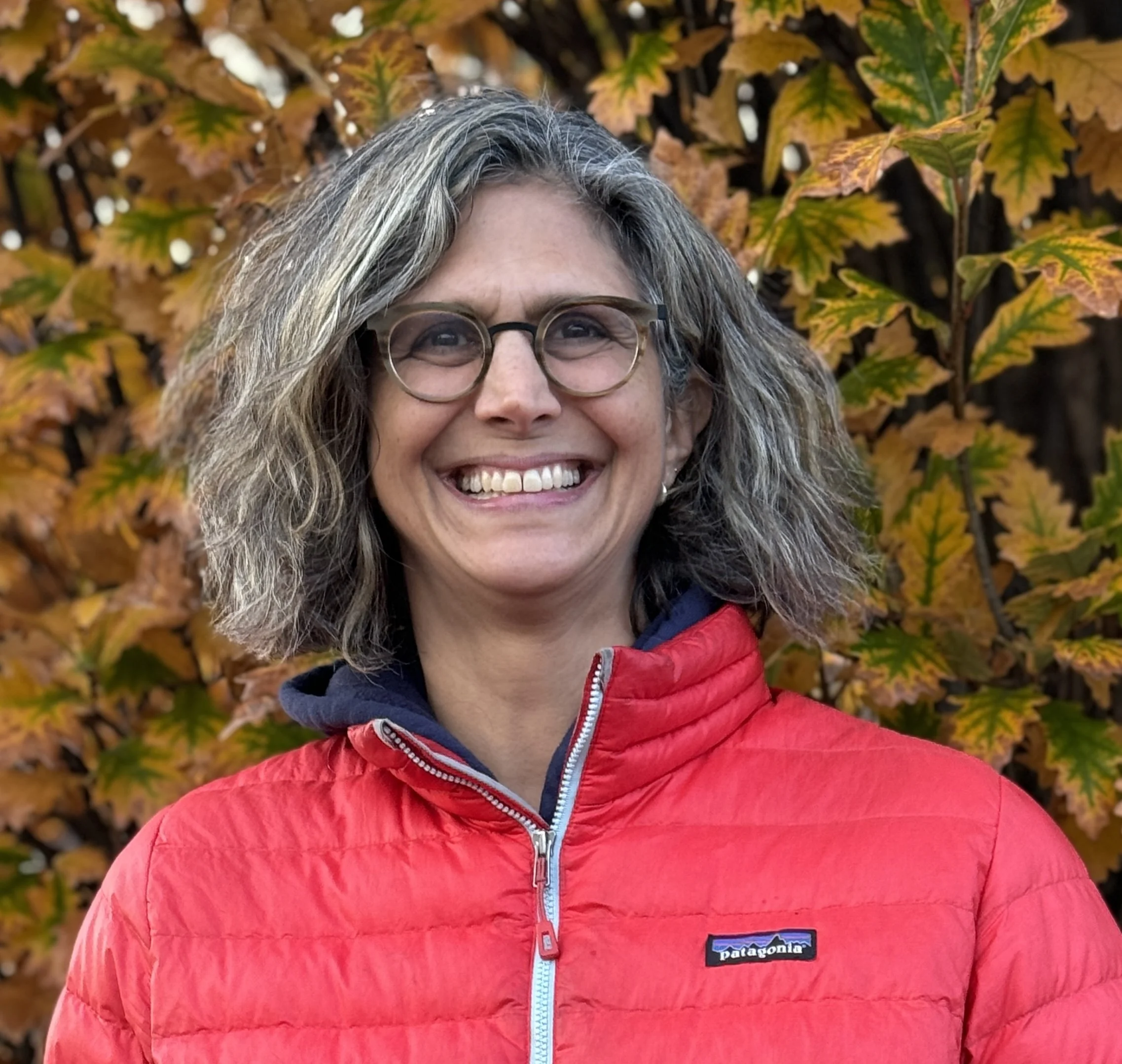 A person with short, wavy gray hair and glasses smiles broadly in front of a backdrop of autumn leaves.