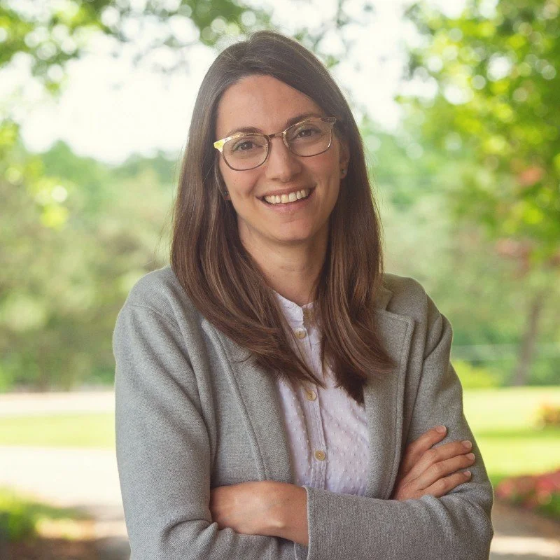 Smiling woman with long brown hair, wearing glasses, a white collared shirt, and a gray blazer, arms crossed, standing in front of a blurred green background.