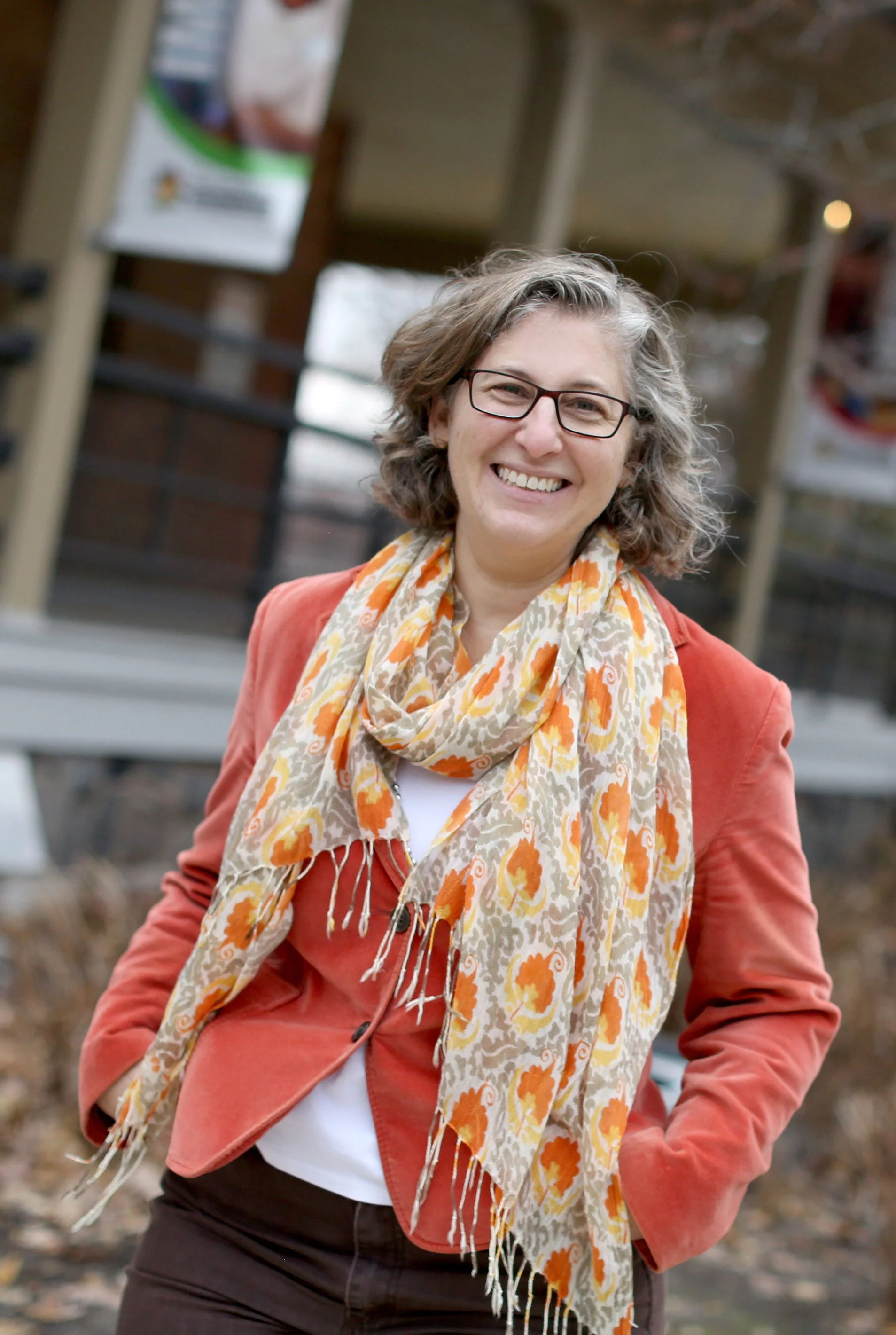 A smiling woman with gray, curly hair and glasses, wearing a rust-colored jacket, a patterned scarf, and brown pants, standing outdoors.